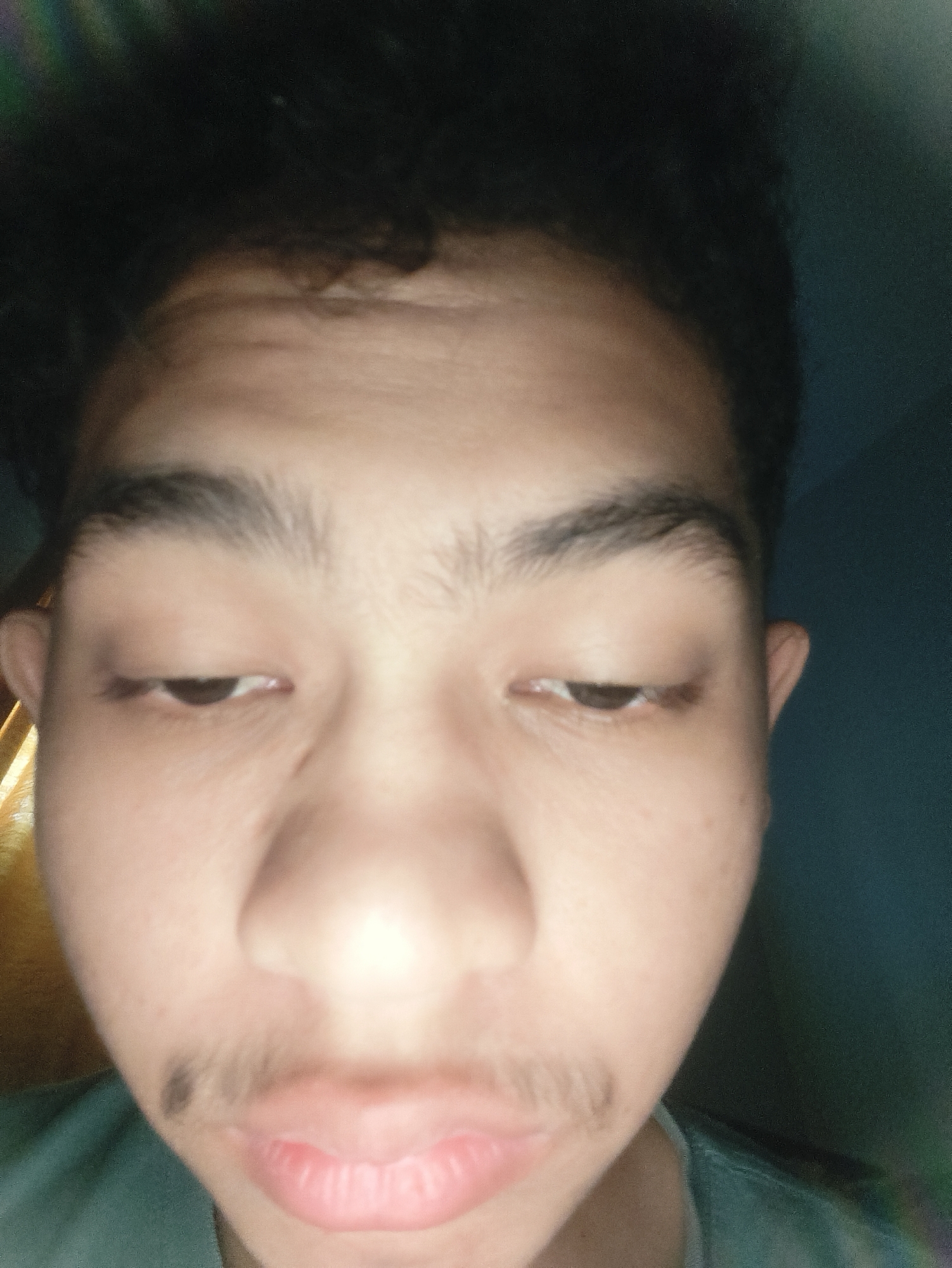 The image displays a close-up, frontal portrait of an individual, with their face dominating the frame. The subject has light-medium skin, dark, tousled hair, and prominent dark eyebrows. Their eyes are partially closed and directed downwards. Sparse facial hair is present, forming a thin mustache above the upper lip and a small dark patch on the subject's left cheek (viewer's right). The mouth is closed, with lips slightly pursed. A collar and shoulder portion of a teal or dark green garment are visible at the bottom left. The background is indistinct and dark, characteristic of an indoor setting. Warmer, yellow-toned ambient light is evident on the left side of the background, while the right side displays a darker, desaturated teal-green tone. The scene is consistent with an indoor environment, reportedly located in the city of Lhokseumawe, Indonesia.