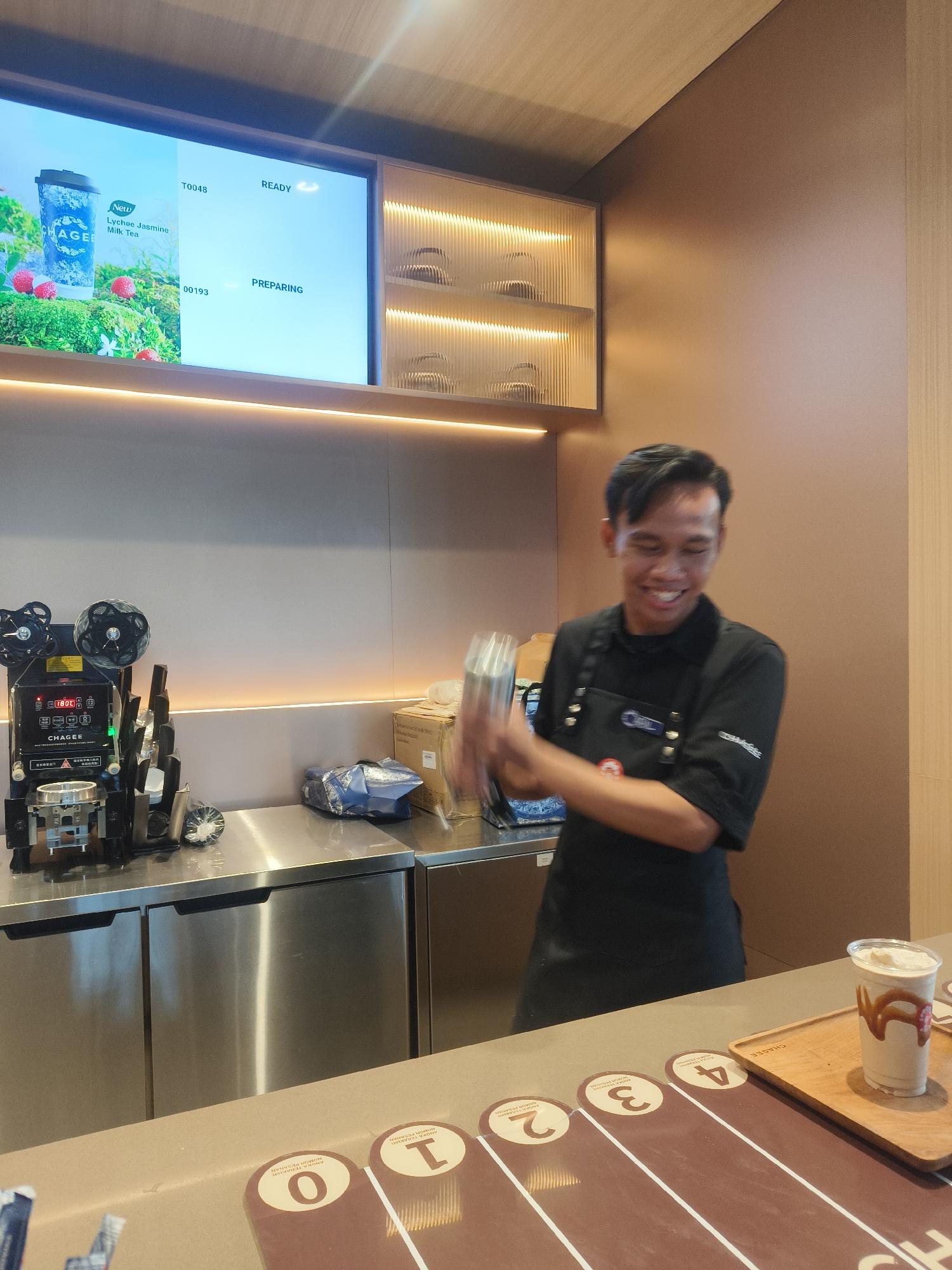 The image shows a young man, likely a barista, working behind a counter in what appears to be a cafe. He is smiling and holding a cocktail shaker, suggesting he is in the process of preparing a drink. He is wearing a black apron over a black shirt. Behind him, a digital screen displays menu items, with "Lychee Jasmine Milk Tea" and "T0048 READY" visible. To his left, there is a drink sealing machine. The counter is made of stainless steel, and the overall environment suggests a modern, clean establishment. In the foreground, a finished drink with caramel drizzle sits on a wooden tray, alongside markers numbered 0, 1, 2, 3, and 4, possibly indicating order positions. The background features a warm-toned wall and decorative lighting, adding to the ambiance. The establishment is located in Kuta Selatan, Indonesia, implied by the context of the cafe.