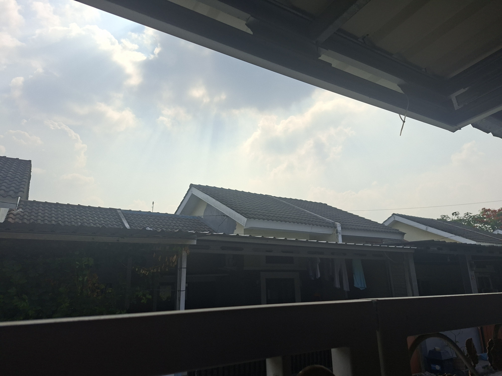 The image captures a residential scene in Ciseeng, Indonesia, viewed from an elevated perspective, possibly a balcony or upper floor, looking out over neighboring houses. The foreground is dominated by a dark, horizontal structure, likely a balcony railing or the underside of an overhang, which frames the lower and upper edges of the shot.
Above the houses, the sky is partly cloudy, with visible sun rays piercing through the clouds, indicating a bright day, likely mid-morning or afternoon. The clouds are a mix of bright white and light gray.
The middle ground features several houses with dark gray or black tiled roofs and light-colored eaves and walls, typical of residential architecture in the region. Many of these houses have carports or covered patio areas in front, under which green foliage is visible on the left. On the right side, clothes are seen hanging to dry on a line under one of these covered structures, suggesting everyday domestic life. A small satellite dish is faintly discernible on one of the roofs.
The overall impression is one of a calm and sunny day in a quiet residential neighborhood. No people or visible text are present in the image.