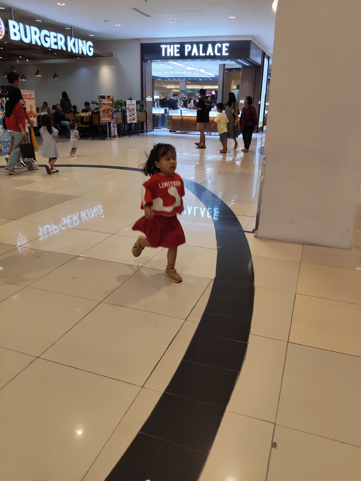 The image captures a scene inside the Sun Plaza mall in Medan, Indonesia. In the foreground, a young girl wearing a red shirt with white lettering and a red skirt is captured in mid-stride, running towards the right. Her hair is dark and slightly windswept. The floor is tiled in a checkered pattern of beige and white, with a black stripe curving through the scene. In the background, the "Burger King" sign is visible on the left, illuminated with white lettering. To the right of Burger King, the entrance to "THE PALACE" store is visible. Several other people are scattered throughout the mall, some walking and others standing. The overall environment appears to be a brightly lit indoor shopping mall.