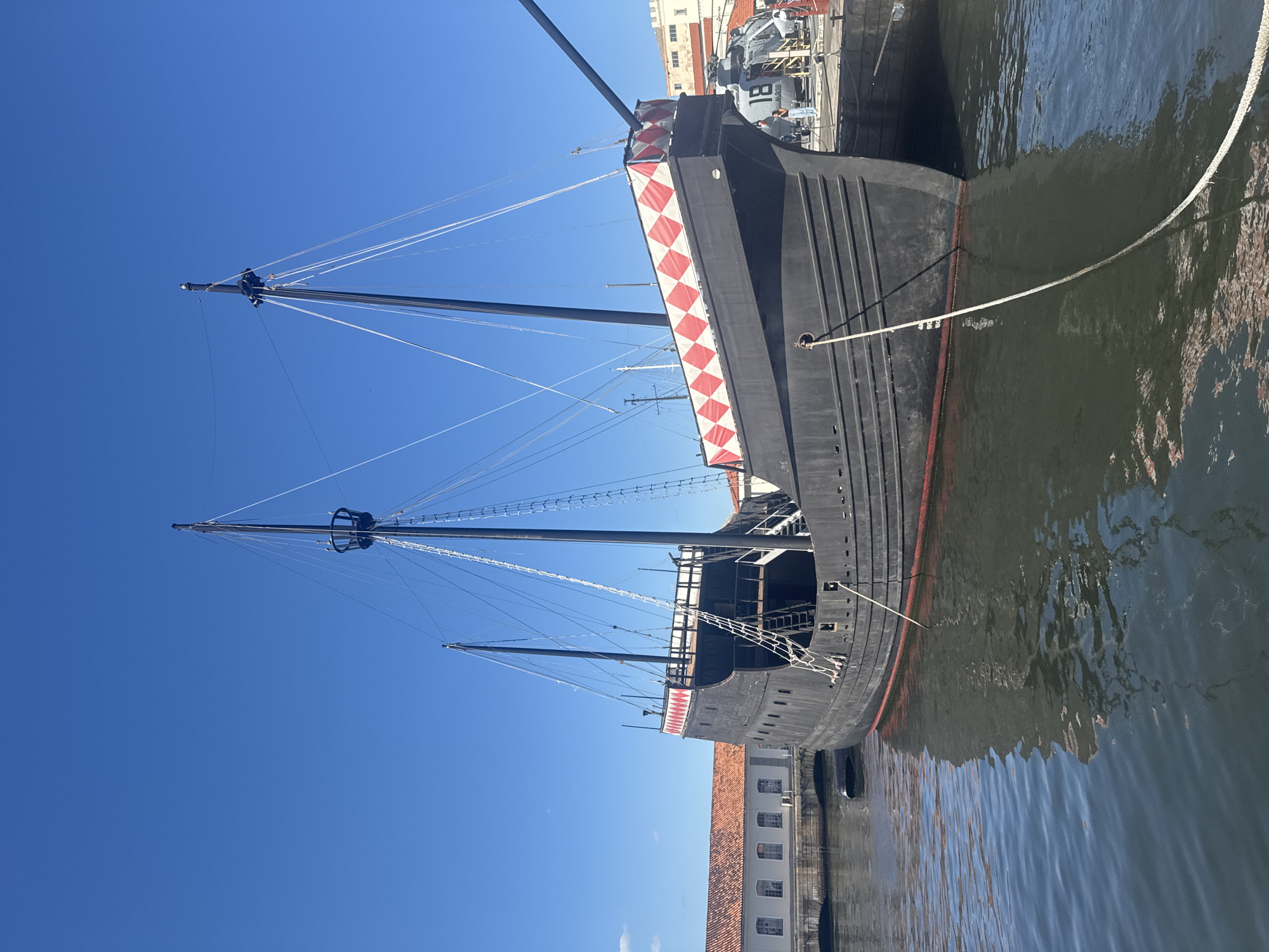 The image shows a black replica of a historic carrack ship docked in a harbor. The ship has two prominent masts with rigging and a decorative red and white diamond pattern along its upper edge. In the background, a building with red tile roofs and white walls is visible along the waterfront. To the right of the ship, a modern helicopter is parked on a dock. A few people are standing near the helicopter, and a thick rope is tied to the ship and extends into the foreground, crossing the water. The scene is set against a clear blue sky. This is the Nau Capitânia museum ship located at Praça XV, Rio de Janeiro, Brazil.