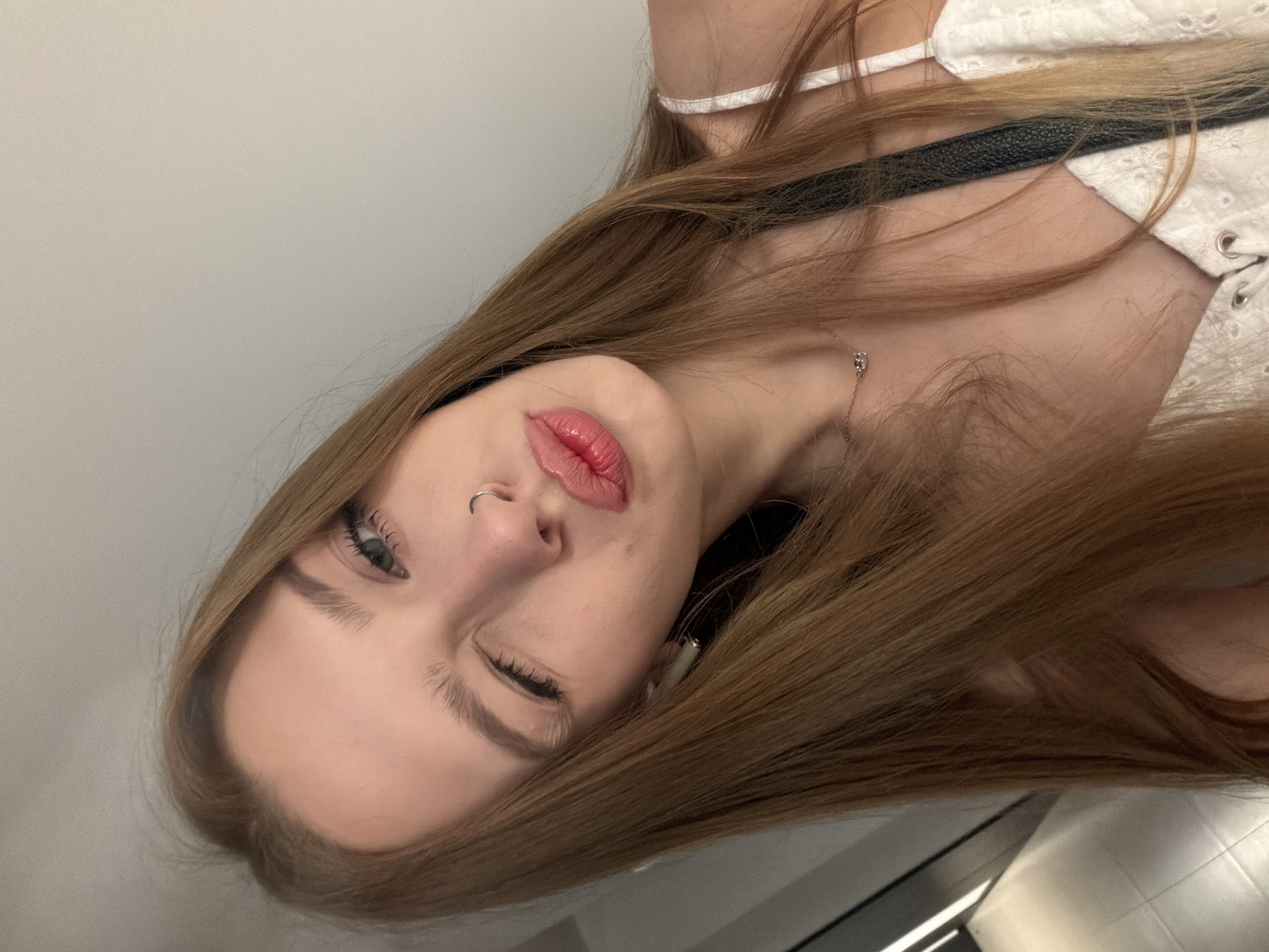 The media file is a close-up, vertically oriented, candid photograph of a young woman. She has long, light brown hair that frames her face, and she is wearing a white top with eyelet detailing and a black strap. A delicate necklace with a small pendant is visible around her neck. Her lips are slightly parted and painted with a glossy pink lipstick. She is winking, and her left eye is open with visible eyelashes and eyebrows. A silver nose ring is apparent on her left nostril. The background is a plain, light-colored wall. The lighting appears to be artificial, suggesting an indoor setting. The image captures a playful or flirtatious moment. There is no visible text.