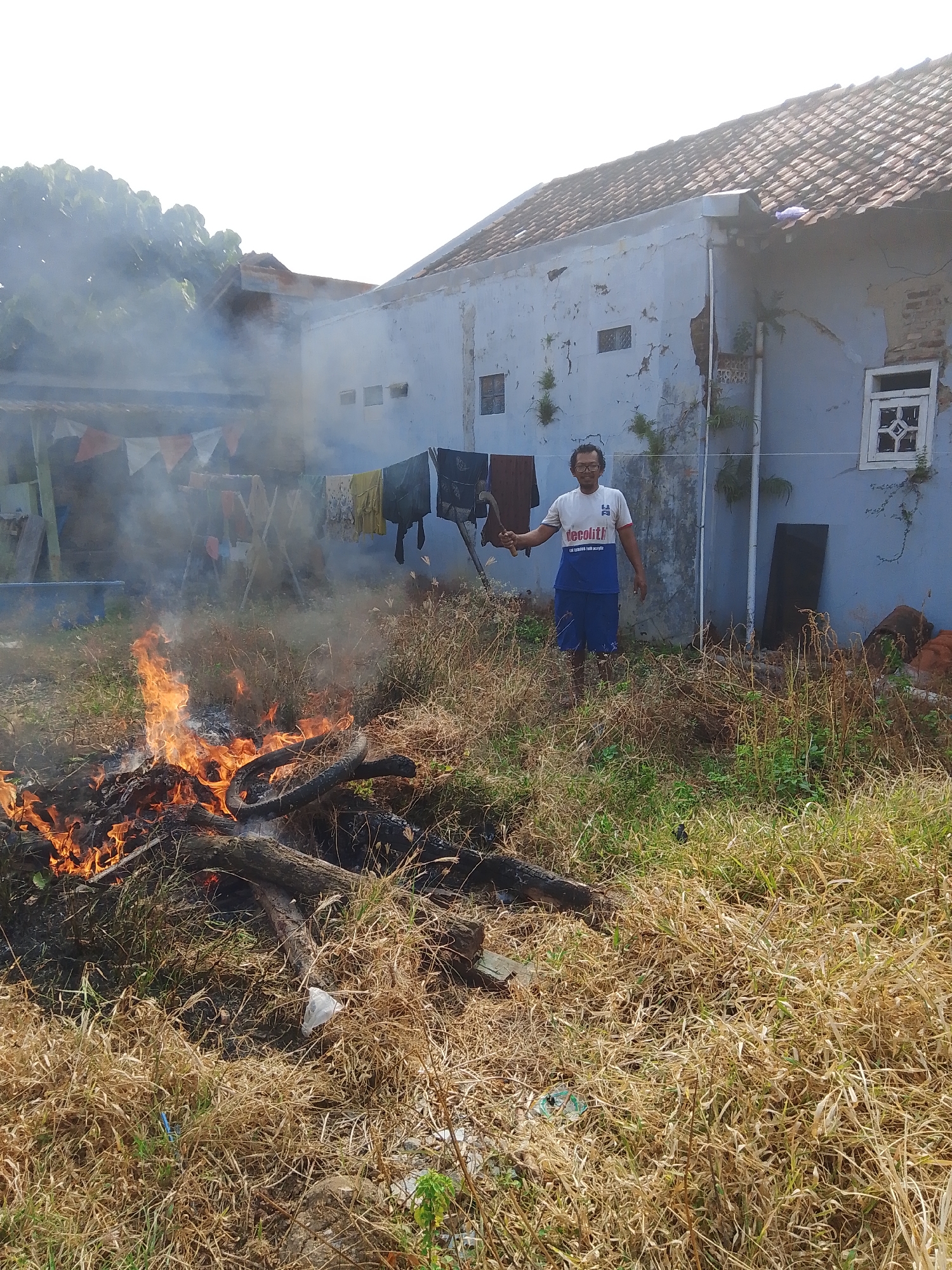 The image depicts an outdoor scene in a residential area, likely in Pekalongan, Indonesia, given the provided context. In the foreground, a bonfire is actively burning, fueled by dried branches and logs, producing significant smoke that drifts upwards and to the left. The ground is covered in dry, overgrown grass and some debris, including a white plastic bag and a deflated tire.
A person, appearing to be a male, stands in the middle ground to the right of the fire. He is wearing glasses, a white t-shirt with blue lettering that reads "decolith" and "cat dinding anti acrilic", and blue shorts. He is holding a tool that resembles a sickle or a machete in his right hand. Behind him is a light blue, somewhat dilapidated building with a tiled roof. Various items of clothing are hung on a clothesline strung between poles in front of the building, partially obscured by the smoke. The building's exterior shows signs of wear, with peeling paint and exposed brickwork in some areas. The overall environment appears to be a backyard or an undeveloped lot adjacent to a dwelling.