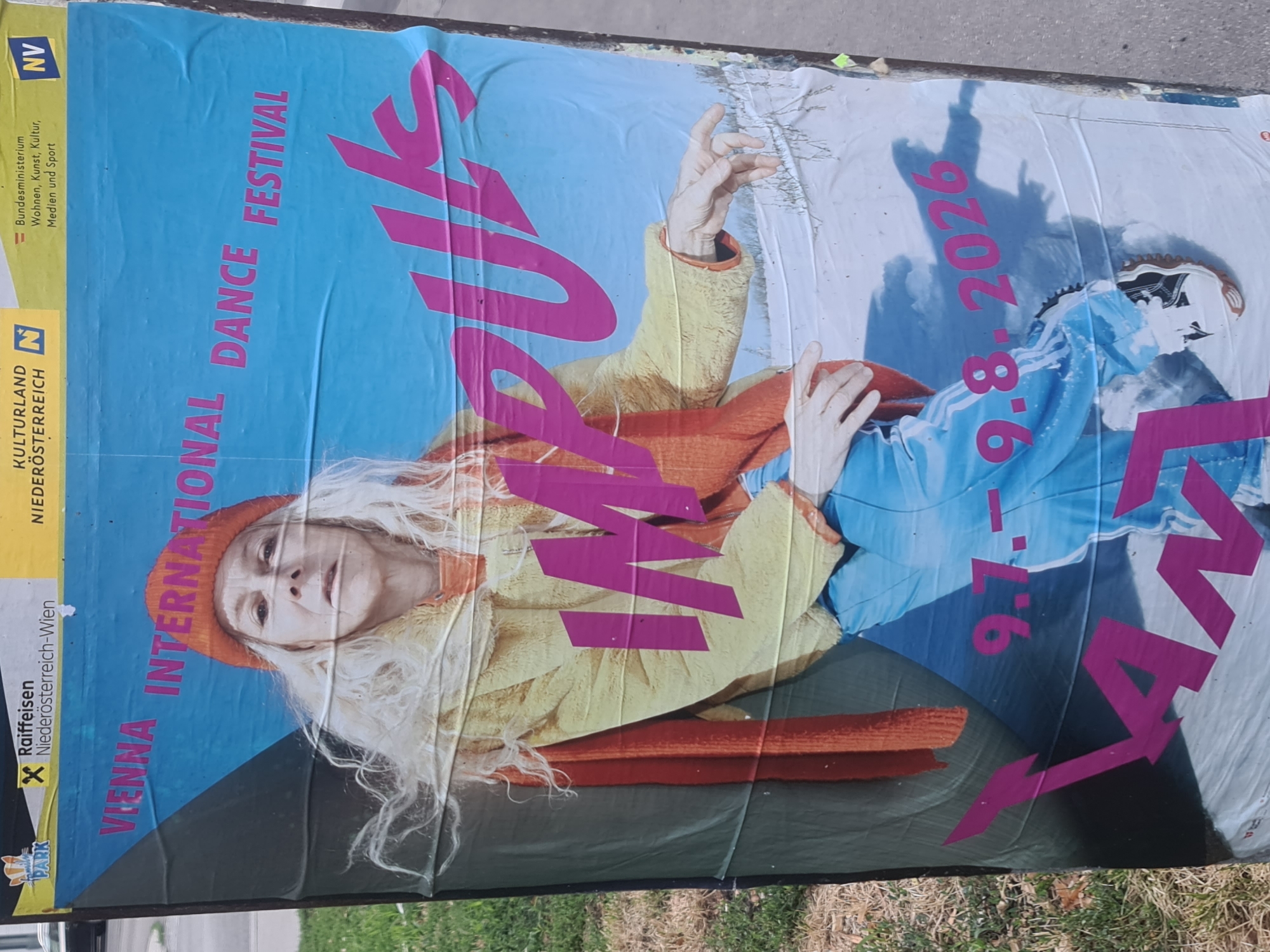 This is a full-frame, eye-level photograph of a torn and weathered poster advertising the "Vienna International Dance Festival" in Vienna, Austria. The poster features a close-up of a woman with long, light blonde hair, wearing a bright yellow fuzzy jacket and a red beanie. She is also wearing blue pants with white stripes. Her expression is neutral. Large pink text on the poster reads "IMPULS" and "VIENNA INTERNATIONAL DANCE FESTIVAL". The dates "9.7. - 9.8. 2026" are also visible in pink text. The background of the poster transitions from a blue sky to a snow-covered landscape. The poster is affixed to a dark pole and is heavily creased and torn in several places, indicating it has been outdoors for some time. The lower portion of the poster appears to be obscured by another poster or material. To the left of the poster, there is a patch of green grass and some dry vegetation. To the right, a concrete sidewalk is visible. The poster is positioned vertically.