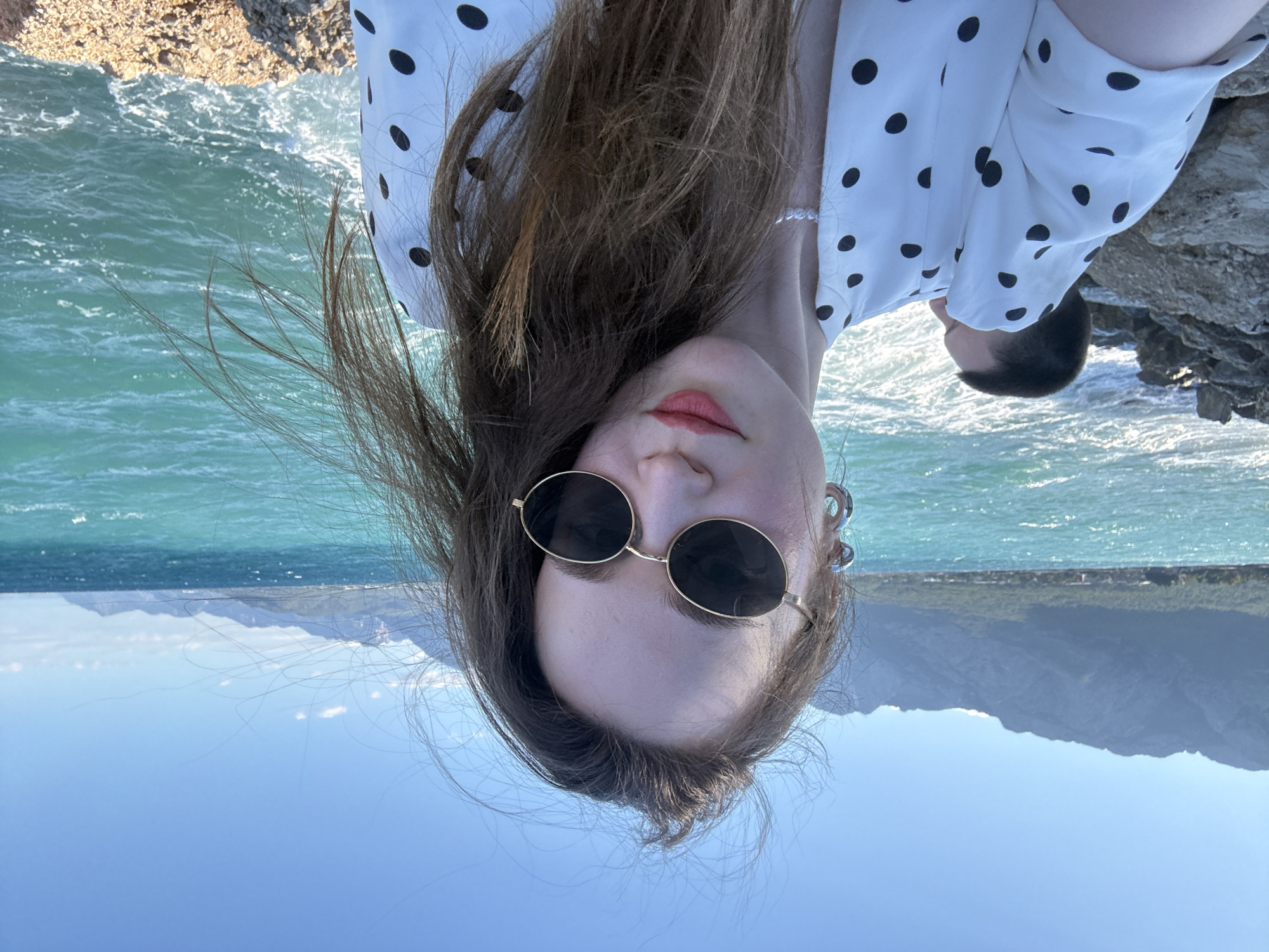 A medium shot selfie shows a woman wearing a white dress with black polka dots, sunglasses, and a pearl necklace. She is standing outdoors with a body of water and mountains in the background. The ocean is turquoise and appears choppy, with waves crashing against a rocky shore. The mountains are jagged and appear to be covered in sparse vegetation. The sky is clear and blue. A man is visible in the background, partially obscured by the woman. The scene appears to be in Kemer, Turkey, a coastal resort town.