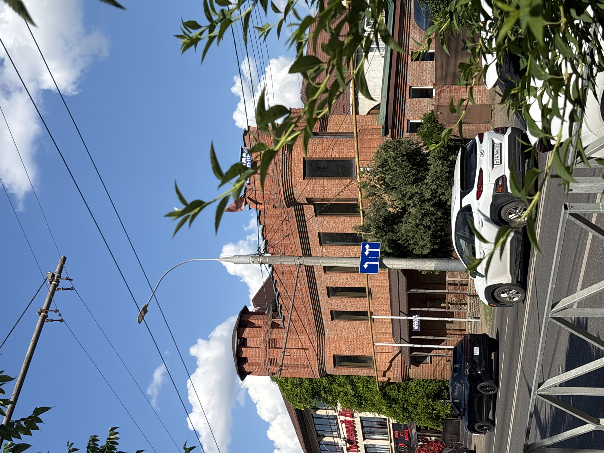 The image shows a street scene in Krasnodar, Russia, on a sunny day with scattered clouds. The perspective is from behind some leafy green branches, looking towards a brick building and a street with cars. The building is multi-story with a distinctive rounded tower-like section. It is covered in some areas with lush green ivy. Several windows are visible, some dark. On the street in front of the building, two cars are parked or driving: a white Hyundai Kona with a license plate that reads "0747EM193" and a black car behind it. There is a traffic sign showing arrows for left and straight directions. Power lines and a lamppost are prominent in the sky. A metal fence or railing is visible in the foreground, on the right side of the image. The overall impression is a bright, urban environment on a clear day. Some text is visible on the building, including "АРЕНА" (ARENA) and other less legible words.