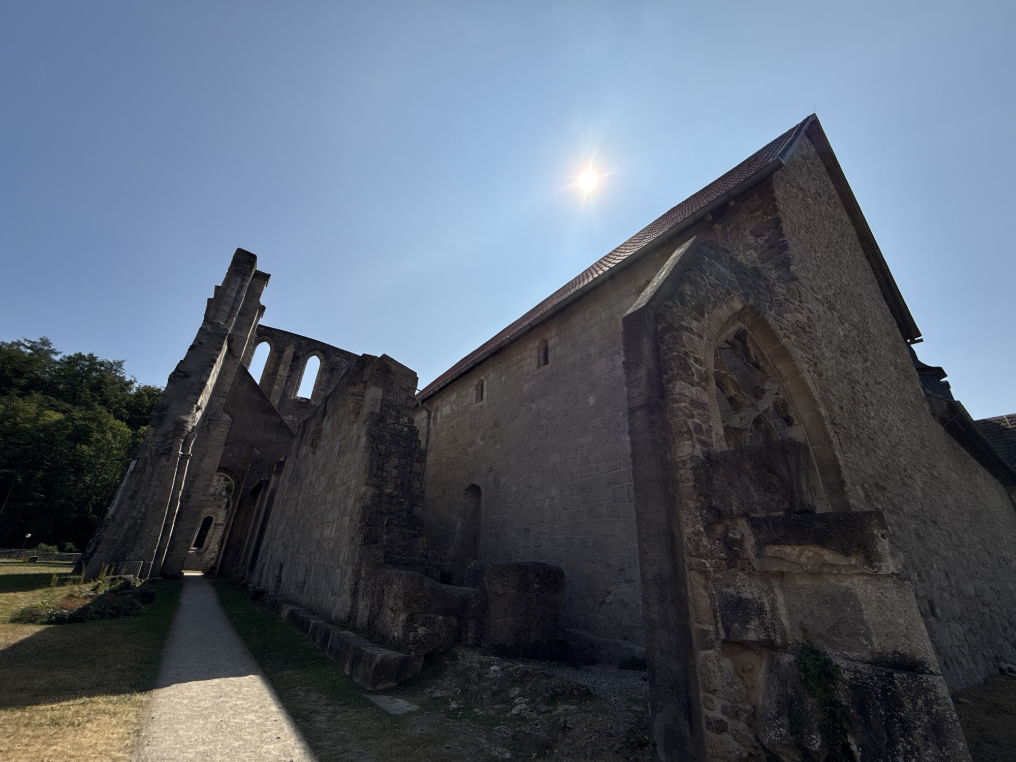 The image shows a low-angle, wide-shot of the ruins of Walkenried Abbey in Walkenried, Germany, on a clear, sunny day. The sun is visible in the upper right quadrant of the sky, casting a bright glare. The abbey ruins are constructed of stone, with portions of the walls still standing, featuring arched windows and a red-tiled roof on one section. A pathway runs along the left side of the ruins, leading into the shadowed interior. Lush green trees are visible in the background to the left. The ground is covered in grass and dry, yellowed patches. No people or animals are present in the image.
