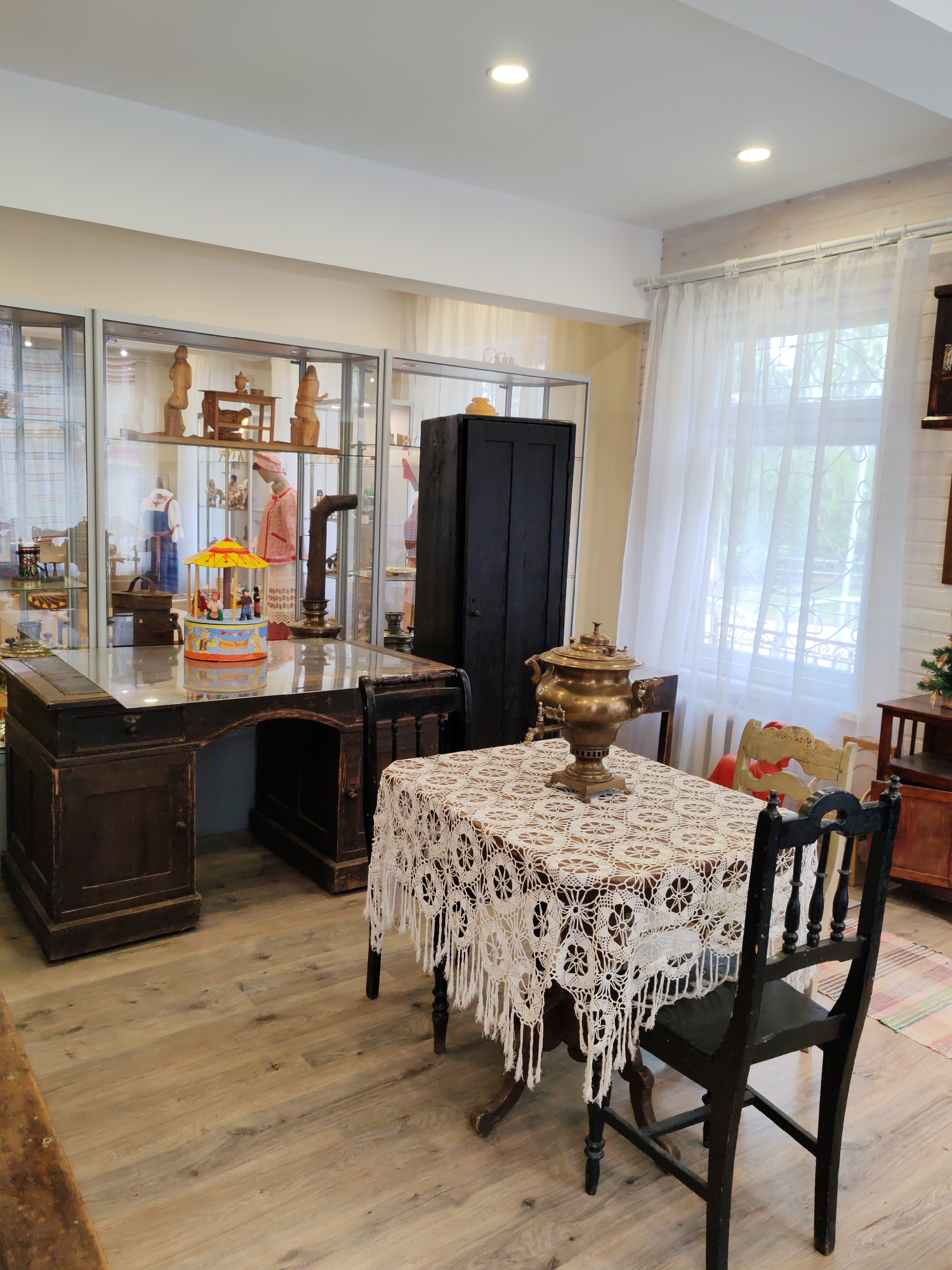 The image depicts an interior scene, likely a museum exhibit, showcasing antique furniture and objects. In the foreground, a dark wooden table is covered with an intricately crocheted white tablecloth. A large, brass samovar sits prominently on the table. A dark, wooden chair with a carved back is positioned at the table. Behind this, a large, dark wooden cabinet stands against a wall. To the left, a dark wooden desk with glass on top has a colorful carousel toy on it. Display cases containing wooden figures and traditional clothing are visible in the background, arranged on shelves. Sheer white curtains cover a window on the right side of the room, allowing natural light to filter in. The flooring appears to be light-colored wood. The overall impression is that of a preserved historical setting, possibly representing a traditional Russian home or exhibit. The location is identified as Vizinga, Russia. There are no people visible in the image.