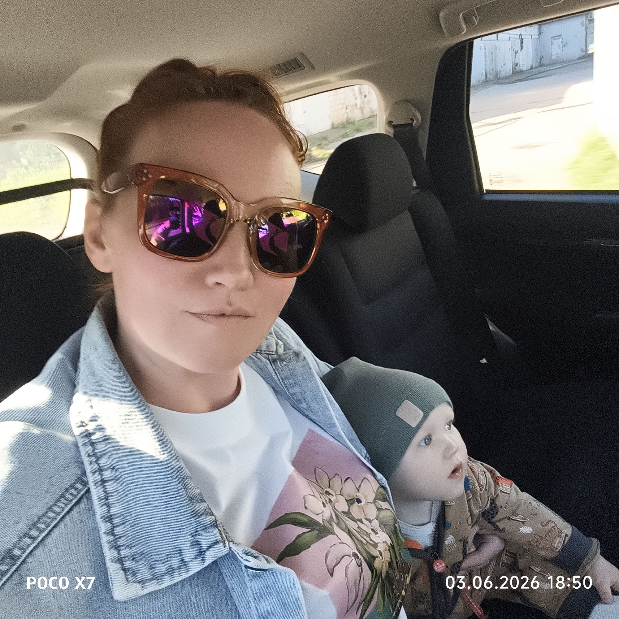The image captures a selfie taken inside a car, featuring a woman and a young child. The woman, positioned closer to the camera, is seen from the chest up, wearing large, orange-framed sunglasses with vibrant magenta and purple reflections. She has reddish-brown hair styled back and is dressed in a light blue denim jacket over a white t-shirt that has a pink and green floral pattern on the front. Her expression is mostly obscured by the sunglasses, but her lips are slightly parted.
Beside her, to the right, is a young child, likely a toddler or baby, wearing a dark green beanie and a light brown jacket with various animal or safari-themed patterns. The child has light blue eyes and is looking intently upwards and to the right, appearing curious or observing something outside the car or above.
The car's interior is visible, with dark seats and door panels. Through the rear side windows, sunlight illuminates the scene, and outside views show what appears to be an urban or industrial environment with light-colored buildings, possibly garages or old structures, and some greenery. The blurred background in one of the windows suggests the car is in motion.
Notable details include the bright reflections in the woman's sunglasses, the detailed patterns on both the woman's t-shirt and the child's jacket, and the contrasting textures of the denim and the smooth car interior.
Visible text includes "POCO X7" in the bottom left corner, indicating the device used, and "03.06.2026 18:50" in the bottom right, providing a date and time stamp, suggesting late afternoon or early evening.