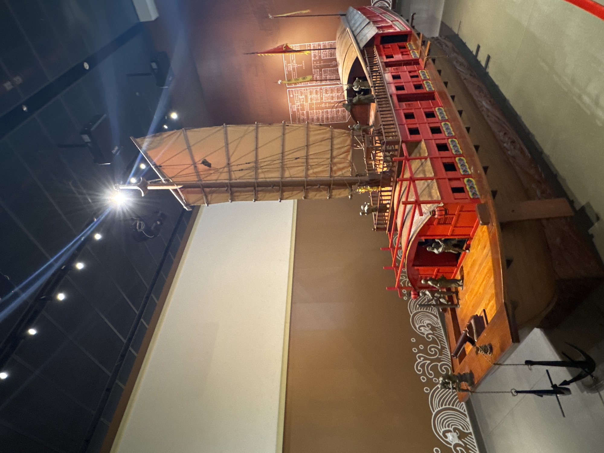 The image shows a large model of a historical Chinese junk ship. The ship is red and brown, with multiple decks and a large sail. There are figures of people on the ship. The model is displayed in a museum or exhibition space, likely in Liwan District, China, based on the style of the ship. The background includes a large blank panel, likely part of the exhibit. The lighting is bright, with a spotlight shining on the sail.