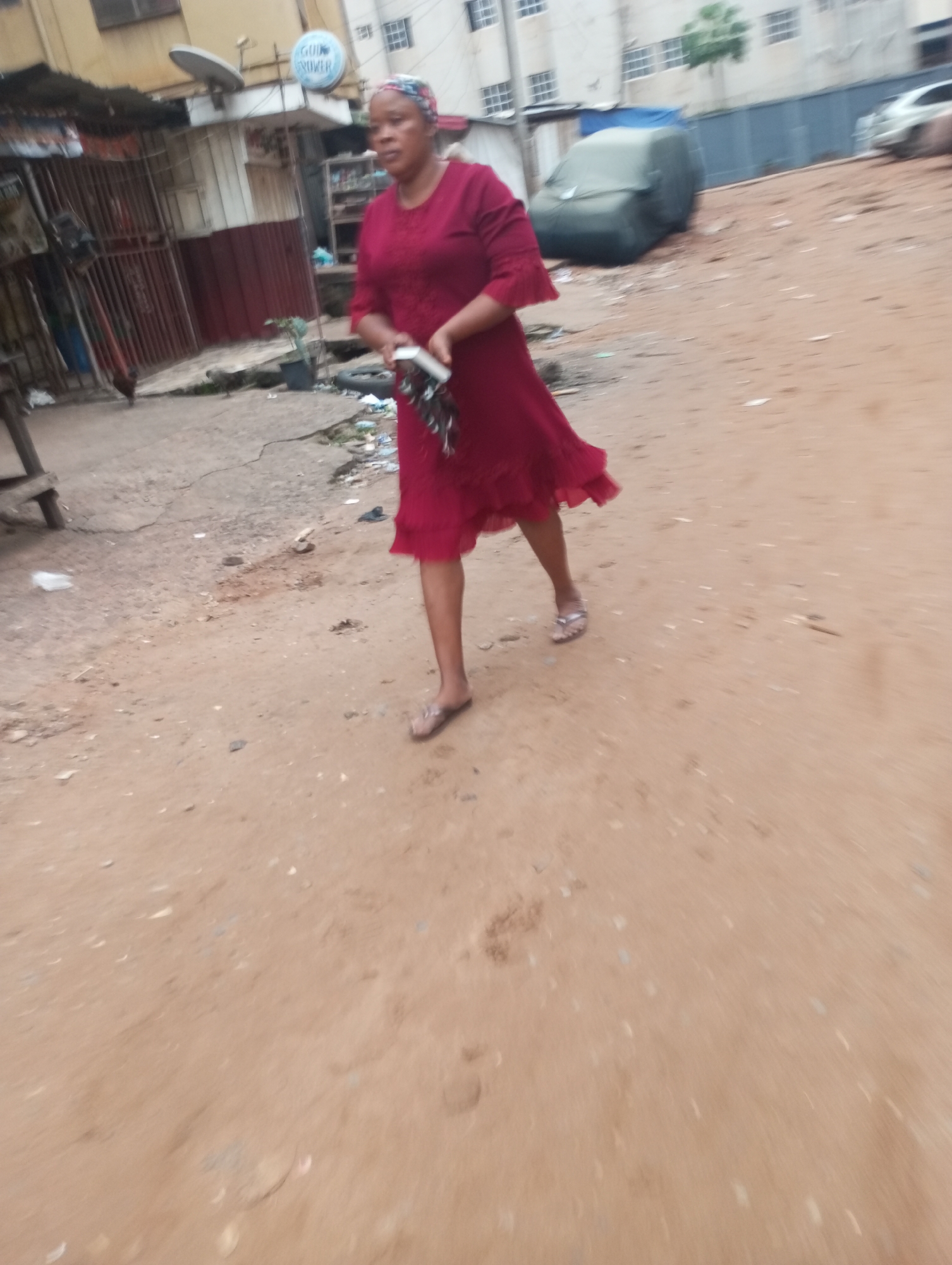 A woman in a red dress walks on a dirt path in what appears to be a market in Onitsha, Nigeria. The woman is wearing a headscarf and holding a white item in her right hand. In the background, there are buildings, a satellite dish, and a vehicle covered with a tarp. The ground is covered with dirt and debris.