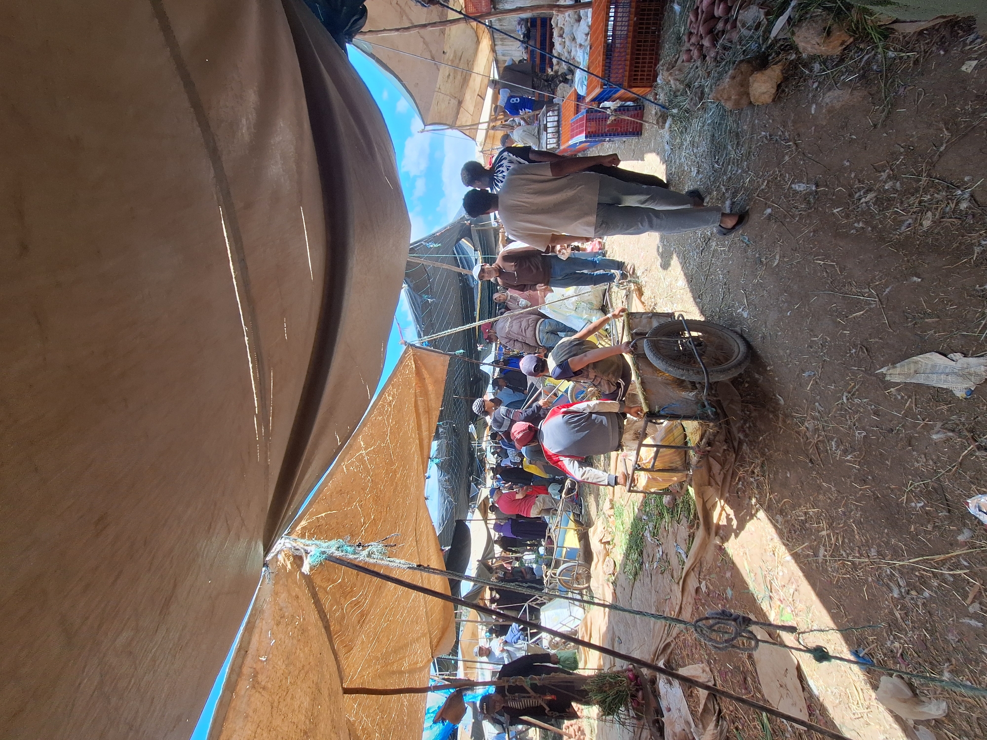 The image depicts a bustling marketplace in Ahrara, Morocco, characterized by rows of makeshift stalls covered with canvas or tarpaulin awnings that create a shaded corridor. The ground is a mixture of dirt and scattered debris. People are densely packed within the corridor, suggesting a crowded shopping environment. Many are wearing casual clothing, with some sporting head coverings. Various individuals are pulling carts loaded with goods. At the end of the corridor, there are stacks of crates filled with produce or merchandise. Overall, the scene conveys a vibrant and active market.