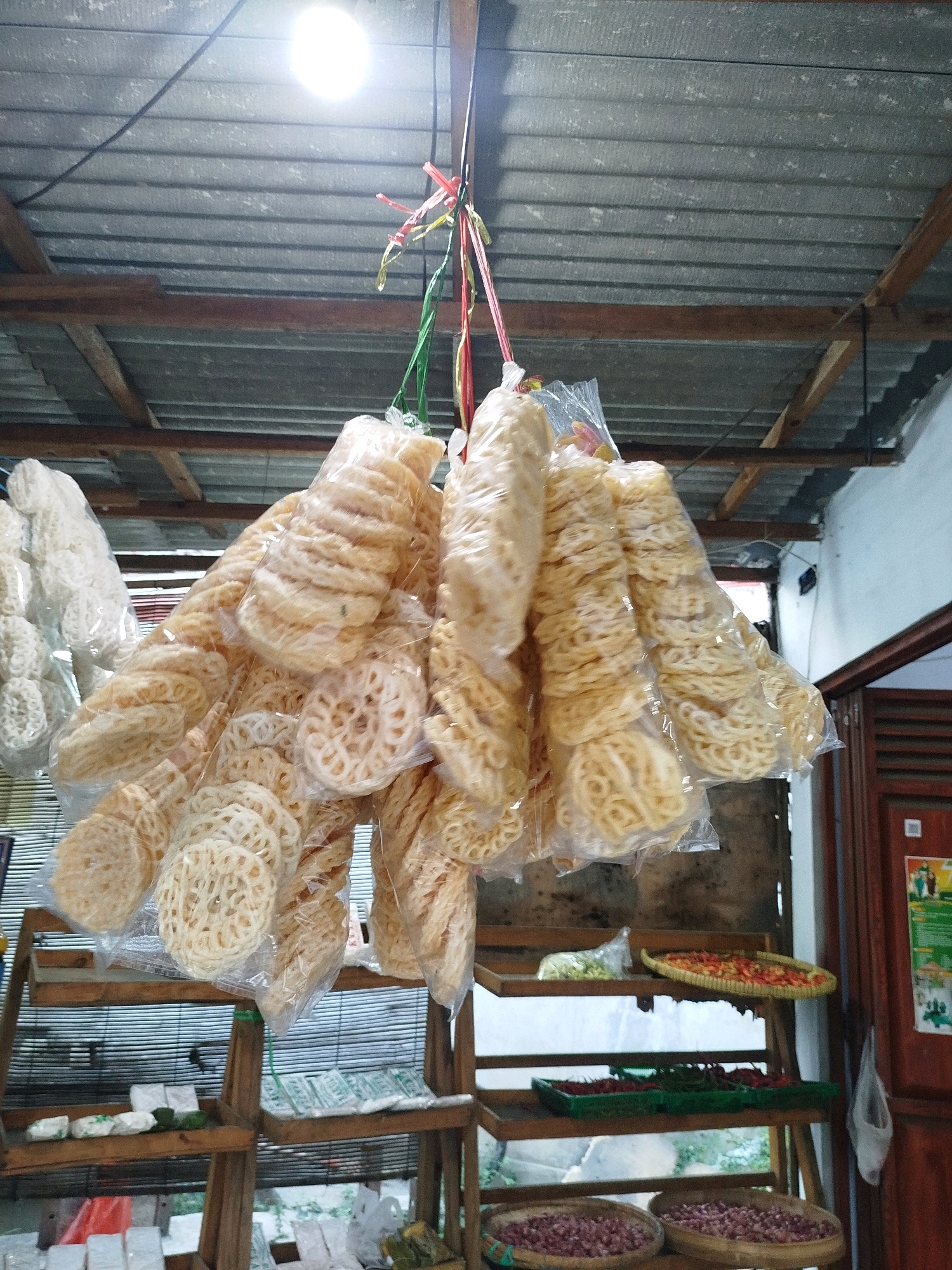 This image depicts a street-side stall in Cekok, Indonesia, selling various types of crackers. The stall features a rustic wooden structure with multiple shelves. Hanging prominently from the corrugated metal roof are numerous clear plastic bags filled with different varieties of light-colored, circular, and ring-shaped crackers. The crackers appear to be made from a flour-based batter and are translucent. Some are flat and round with intricate cut-outs, while others are stacked in a more cylindrical or coiled fashion.
The shelves below display additional goods. One shelf holds what appear to be small, wrapped food items or packets. Another shelf contains baskets filled with what look like red chilies and possibly other vegetables, alongside a tray of red, small, possibly dried food items. The bottom shelves contain two large woven baskets filled with what appear to be red onions or shallots. The background shows a partially visible wooden door and a wall, suggesting an outdoor or semi-outdoor setting. The overall impression is that of a traditional market stall offering fresh produce and snacks.