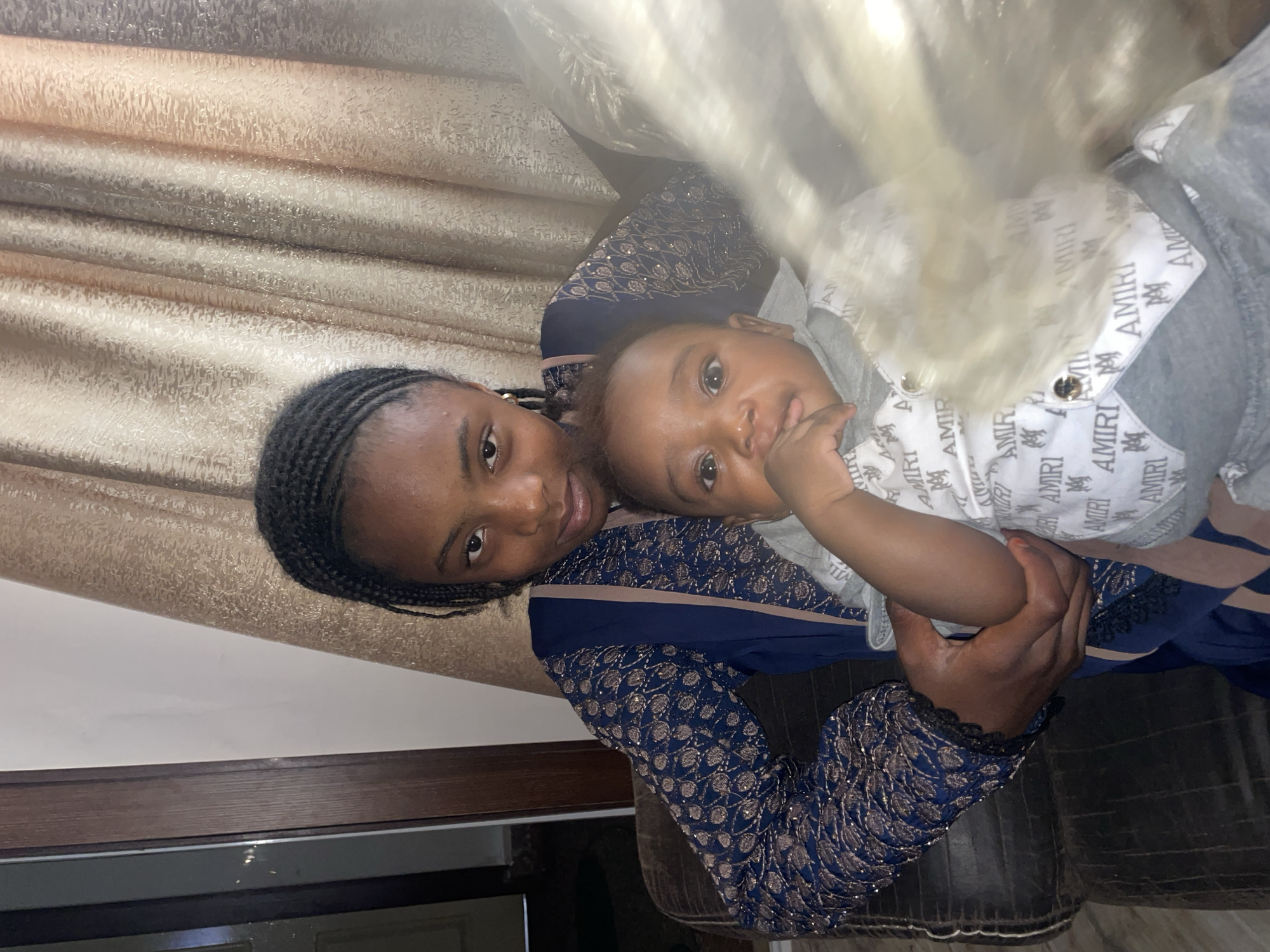 A woman with braided hair is holding a baby in her arms. The baby is wearing a grey and white outfit with the brand name "AMIRI" printed on it. The woman is wearing a blue and gold patterned dress. They are positioned in front of a patterned wall or curtain. The location is difficult to determine from the image, though the query suggests it is in the city of Jos, Nigeria.