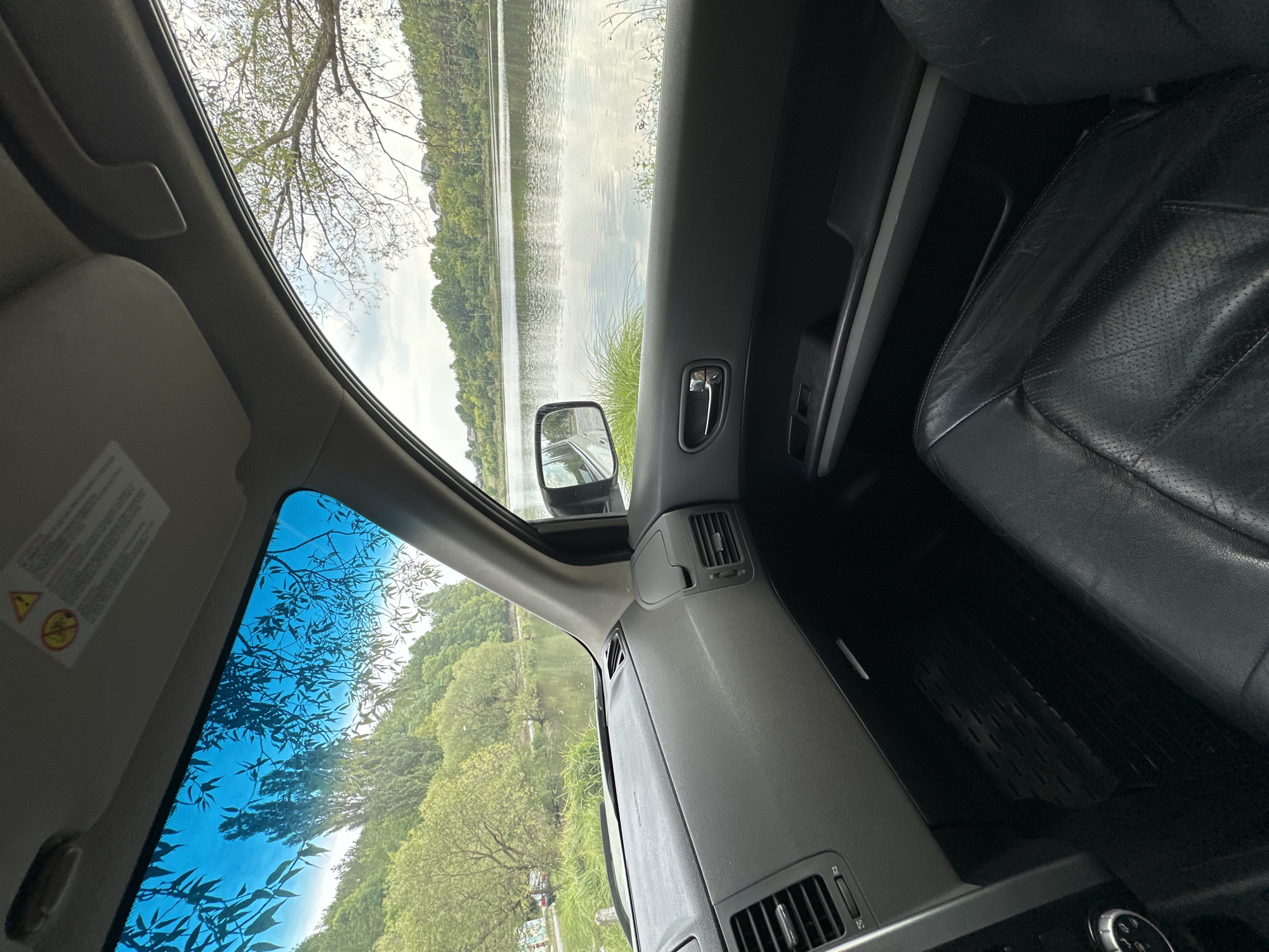 The image shows the interior of a car parked near a body of water, likely a lake or river, with trees and greenery on the opposite bank. The view is from the driver's or passenger's seat, looking out the windshield and side window. The dashboard, car door, and a portion of the leather car seat are visible. The exterior scene depicts a natural landscape with calm water reflecting the sky and trees. No people are present in the image. The location is identified as Makiv, Ukraine.