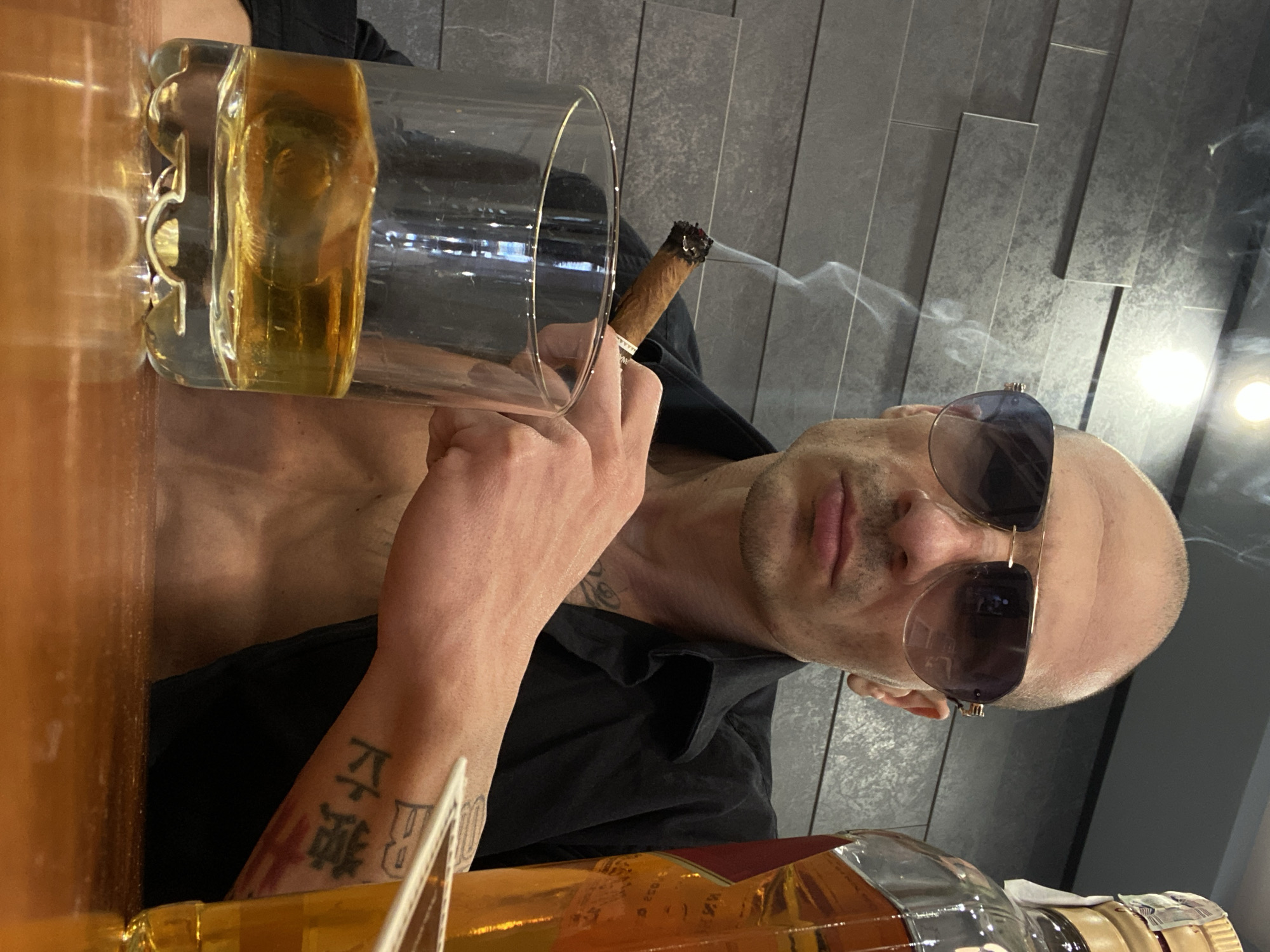 A bald man wearing sunglasses is smoking a cigar and holding a glass of amber liquid, likely whiskey. The man is also near a bottle of amber liquor. He has tattoos on his left arm. The background shows a wall with gray, vertical panels. The photo appears to be taken from an angle looking up at the subject. The location in the city of Pleven, Bulgaria, cannot be determined from the image.