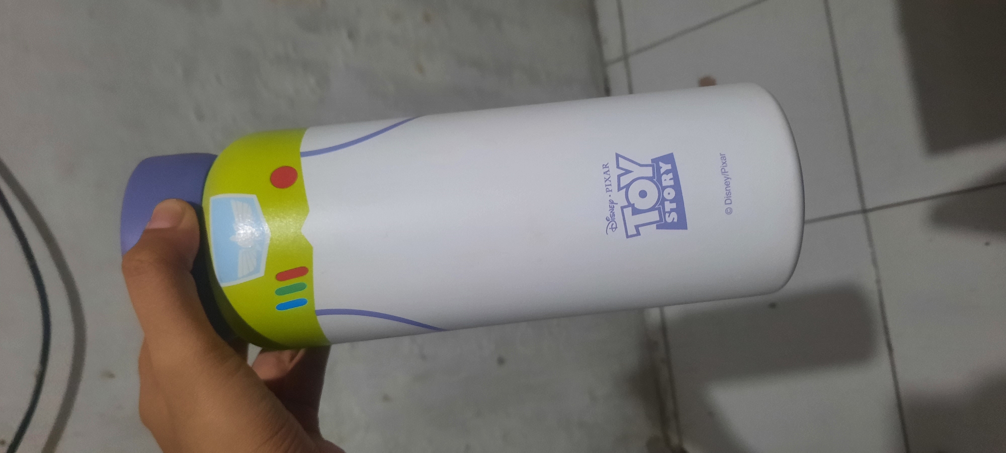 A hand holds a white, cylindrical water bottle designed to resemble Buzz Lightyear from Toy Story. The top of the bottle has a purple cap and a lime green section with blue and red buttons, a white emblem with wings, and a red circular accent. The bottle's body is white with a blue line design. Near the bottom of the bottle, the "Disney PIXAR TOY STORY" logo is visible in purple lettering, along with a smaller copyright notice for "Disney/Pixar". The background is a textured, light-colored wall. This image was taken indoors in North Jakarta, Indonesia.