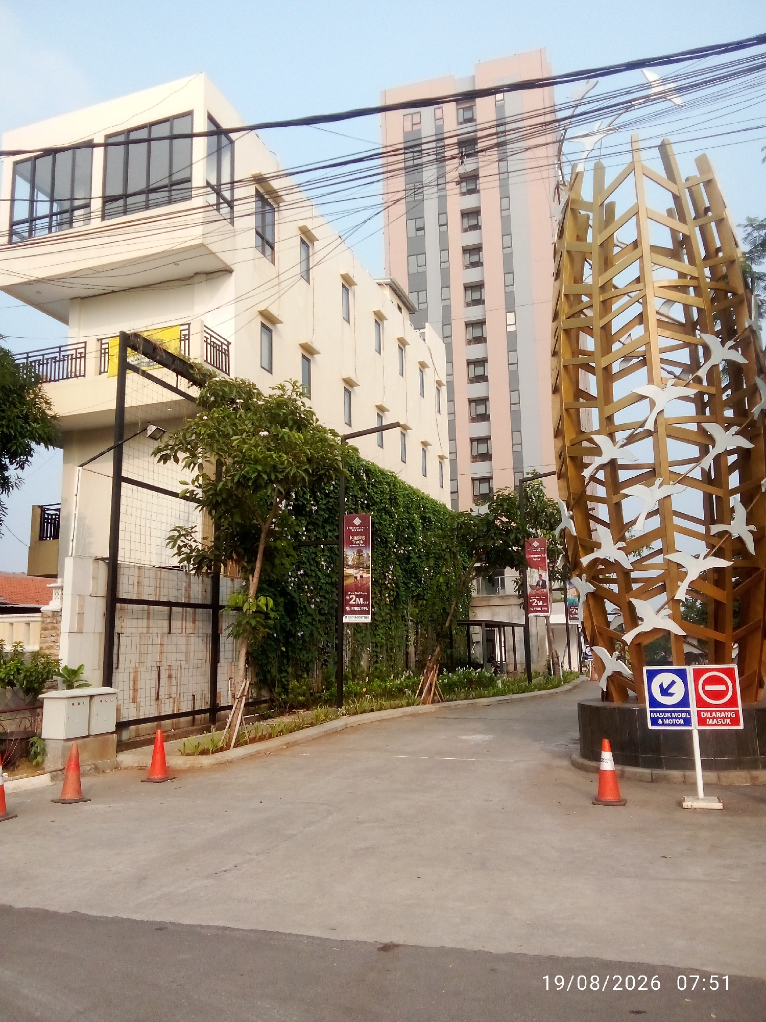 The image depicts a street scene in East Jakarta, Indonesia, likely near a commercial or residential development. In the foreground, a paved road leads towards a large, abstract sculpture made of golden wooden beams and white bird cutouts. To the left of the sculpture, a tall, light-colored building with multiple floors and dark-trimmed windows is visible. Behind this building, a taller, multi-story building with a pink and gray facade stands. A security gate with a black metal fence is present to the left of the road, with lush green foliage and vines growing along it. Several orange traffic cones are placed on the road, with one in the immediate foreground and two further back. To the right of the sculpture, two signs are mounted on a pole: one with a blue downward arrow indicating "MASUK MOBIL & MOTOR" (Car & Motorcycle Entry) and another with a red circle and diagonal line indicating "DILARANG MASUK" (No Entry). The sky is clear and blue. A date and time stamp "19/08/2026 07:51" is visible in the lower right corner.