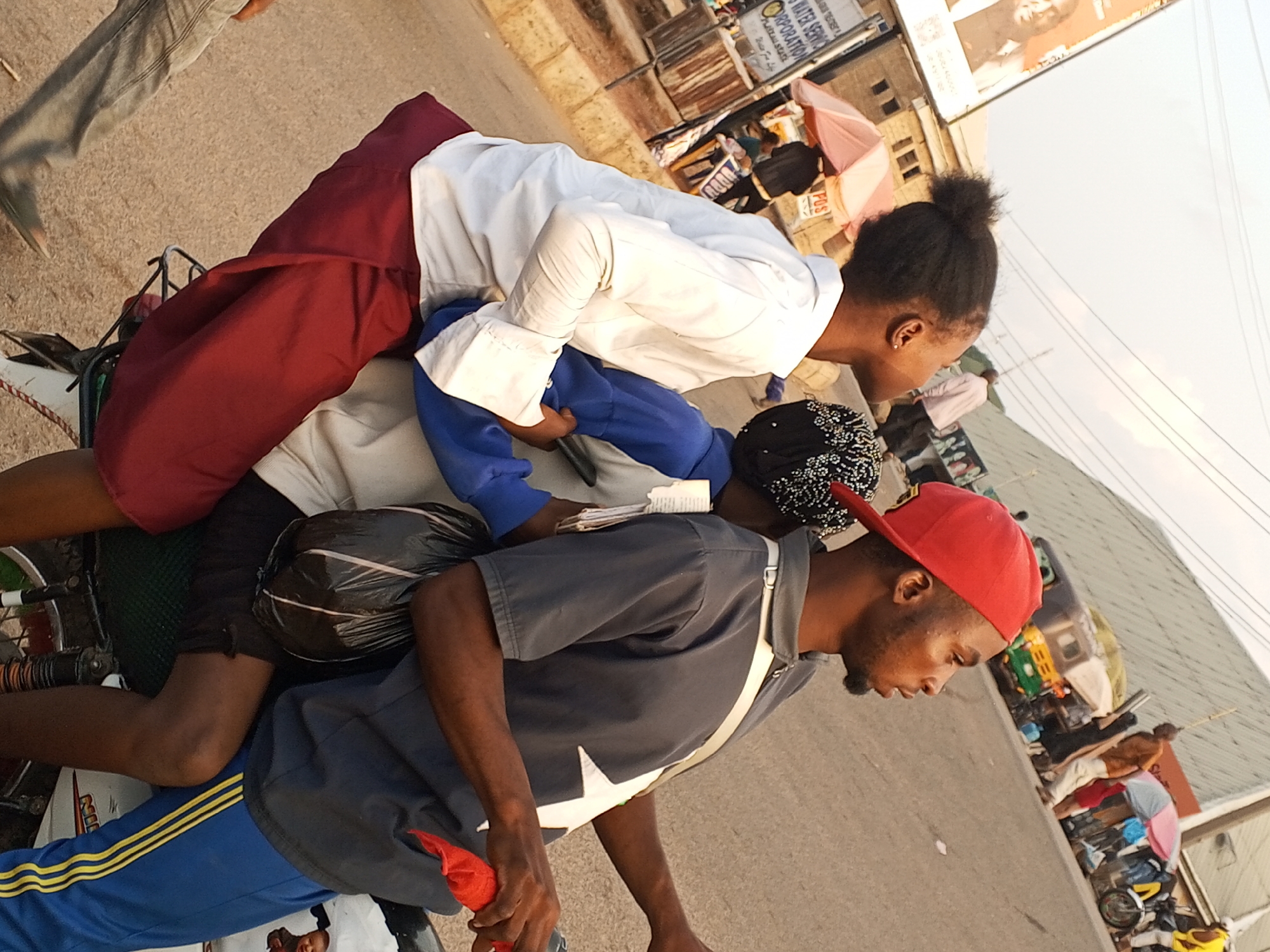 The image depicts a crowded scene in Gwong, Nigeria, with multiple individuals on a motorcycle. The rider wears a red cap and a gray shirt with a white star emblem and blue sweatpants with yellow stripes. Three passengers are precariously positioned on the motorcycle. One wears a white shirt and maroon pants, another wears a blue shirt, and a third is behind them with a patterned head covering. People and vehicles, including auto-rickshaws, are visible in the background, suggesting a busy urban environment. A billboard is seen in the background.
