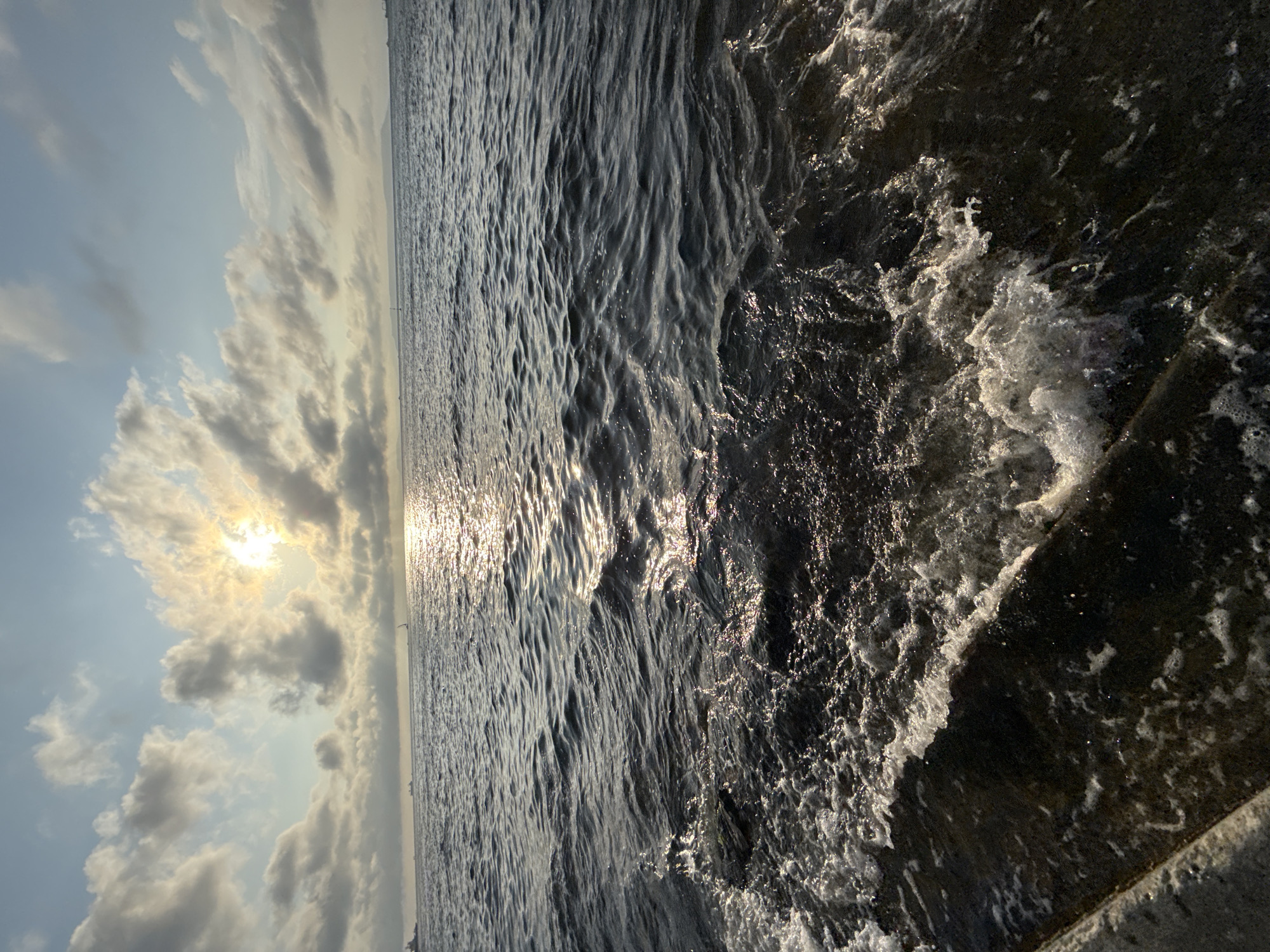 A wide-angle, low-angle shot captures a body of water with choppy waves lapping against a rocky shore. The sun, partially obscured by scattered clouds, casts a shimmering reflection on the water's surface. In the distance, a faint horizon line separates the sea from a pale blue sky. Several ships and possibly a buoy are visible on the water. The foreground features a concrete structure, possibly a seawall or pier, with water breaking over it and creating foam. The overall scene depicts a seascape at sunset or sunrise. The image does not contain identifiable landmarks that would confirm its location in Vladivostok, Russia.
