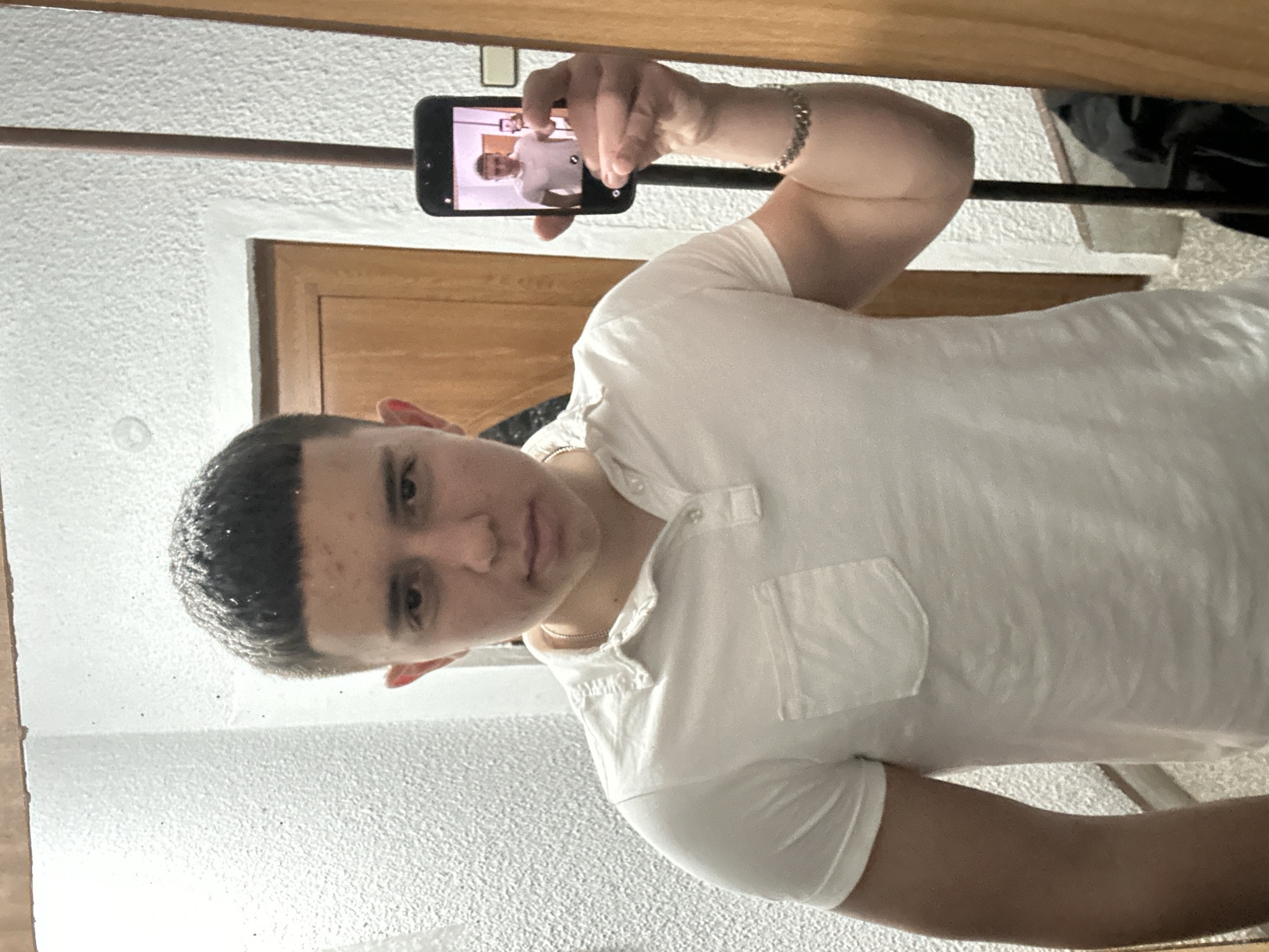 A young man is taking a selfie in a mirror using his smartphone. The reflection in the smartphone's screen shows another image of himself, dressed similarly. He is wearing a white polo shirt and a silver bracelet. The background includes a wooden door frame and a textured white wall, potentially indicating an indoor setting. The image was taken in Zlatograd, Bulgaria.