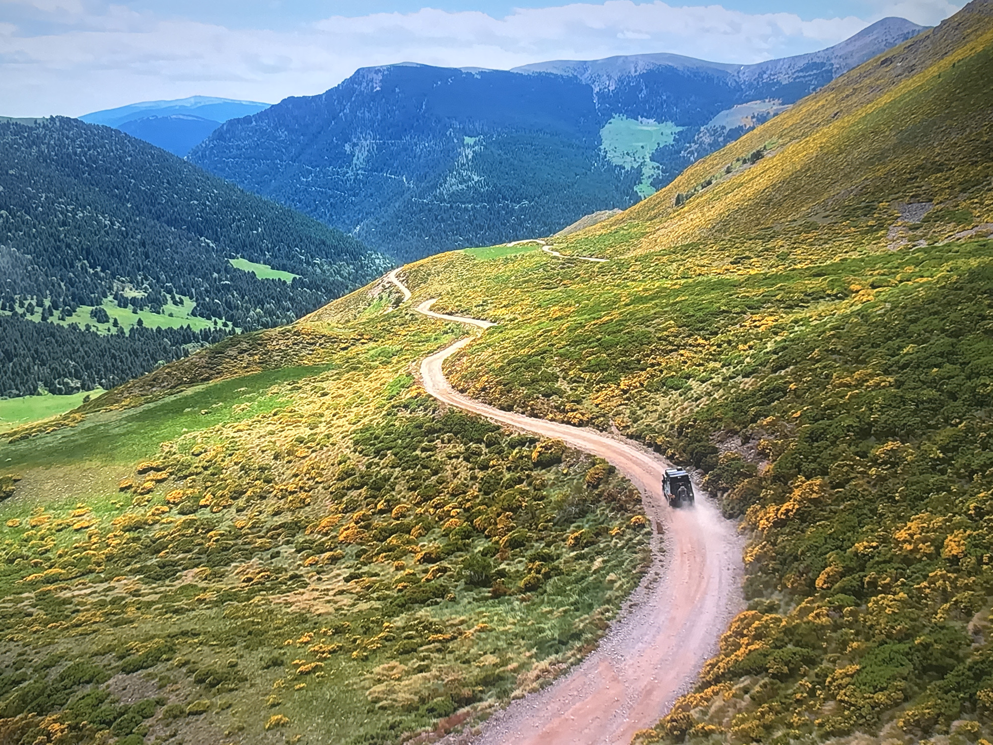 An aerial, eye-level shot captures a winding dirt road cutting through a lush, mountainous landscape. The road is a light brown, appearing dusty as a dark-colored off-road vehicle drives along it, kicking up a trail of dust. The slopes on either side of the road are covered in a vibrant tapestry of green grass and abundant yellow wildflowers, creating a striking contrast. In the background, rolling hills transition into larger, more rugged mountains. The sky above is a pale blue with scattered white clouds. The overall scene depicts a remote, natural environment, likely a rural or undeveloped area.