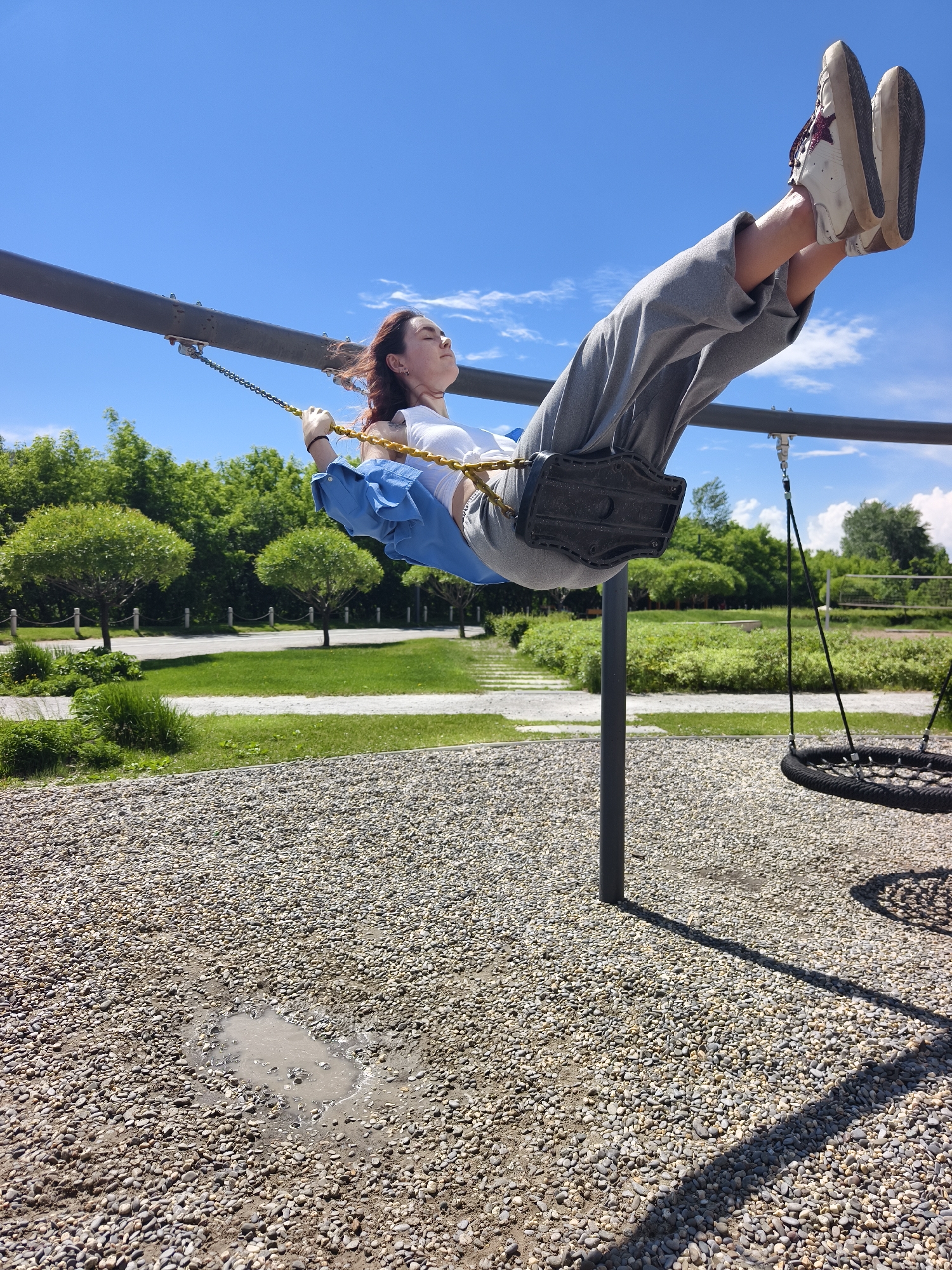 The image depicts a person, appearing to be a young adult with reddish-brown hair, swinging high on a playground swing on a bright, sunny day. They are wearing a white top, a blue long-sleeved shirt tied around their waist, grey sweatpants, and light-colored sneakers with a dark star detail. Their head is tilted back with eyes closed, suggesting a moment of joy or relaxation as they are elevated.
The scene is set in an outdoor park or playground, likely in Tyumen, Russia, as indicated by the context. The ground beneath the swings is covered in light-colored gravel, and a small, muddy puddle is visible directly below the person, indicating recent rain or wet conditions. In the background, verdant green trees and bushes line a paved pathway, enhancing the natural park setting. Another swing, a circular basket-style design, is partially visible to the right. The sky is a clear, vibrant blue with a few scattered white clouds, and bright sunlight casts distinct shadows, confirming it is daytime. There is no discernible text visible in the image. The overall impression is one of carefree outdoor recreation.