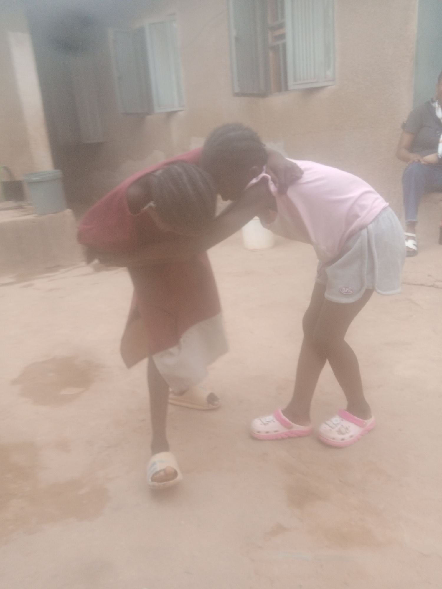 Two children are engaged in what appears to be playful wrestling on a concrete surface. One child wears a maroon shirt with white shorts and light-colored sandals. The other child has on a pink shirt, grey shorts, and pink and white clogs. They are standing close together, with their arms around each other.
Behind them is a building with windows and a seated person. A bucket can be seen to the left of the children. The environment looks to be in Sabon Tasha, Nigeria, based on visual and cultural context. The image is hazy and somewhat out of focus.