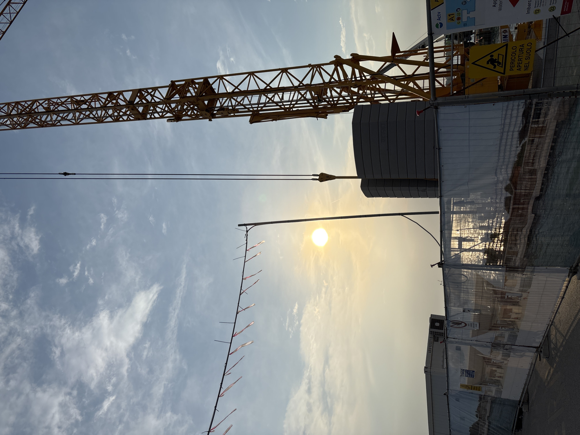 The image depicts a construction site in Punta Sabbioni, Italy, under a bright sun. A yellow crane stretches across the top portion of the frame, partially obscuring the blue sky with scattered clouds. A safety fence, featuring a hazard sign in Italian ("PERICOLO APERTURA NEL SUOLO" - Danger Open Ground), surrounds the perimeter. A utility pole with red and white pennants runs vertically along the left side of the image. Other signs and construction related equipment can be seen in the background.