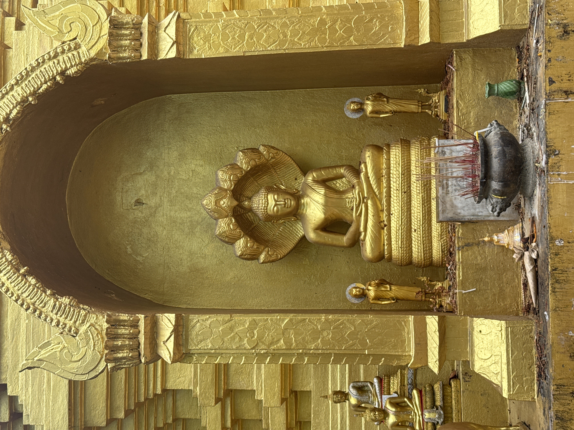 The image depicts a golden statue of a reclining Buddha, shielded by a multi-headed cobra, housed within a golden alcove. Two small golden Buddha figures stand above and below. To the right, there's a dark gray sculpture of a creature, potentially a turtle, with incense sticks in front of it. The entire structure, made of golden material, appears to be part of a Buddhist temple or shrine. The location is Ban Sai Kham, Thailand.