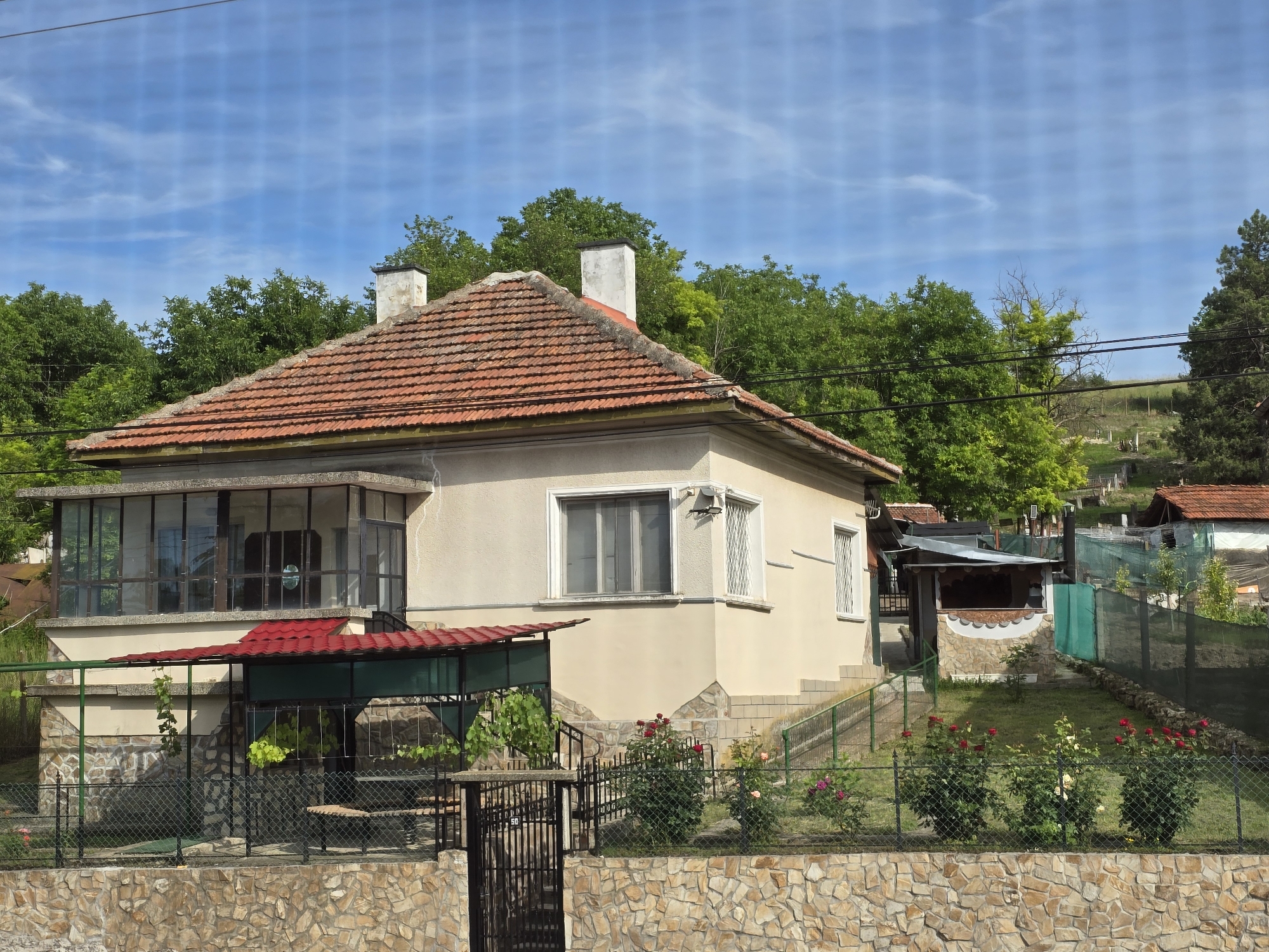 The image shows a light-colored, single-story house with a red tile roof and two chimneys. The house has a sunroom with large glass windows on the left side and a covered patio with a red roof in front of it. There are rose bushes and a fence in front of the house, and a stone wall at the very front of the property. A small outbuilding with a wooden roof is visible to the right of the main house, and further in the background, there are more buildings, trees, and a rolling hill. The sky is blue with some wispy clouds. The scene appears to be in a rural or suburban area in Galatin, Bulgaria. There are no people visible in the image.