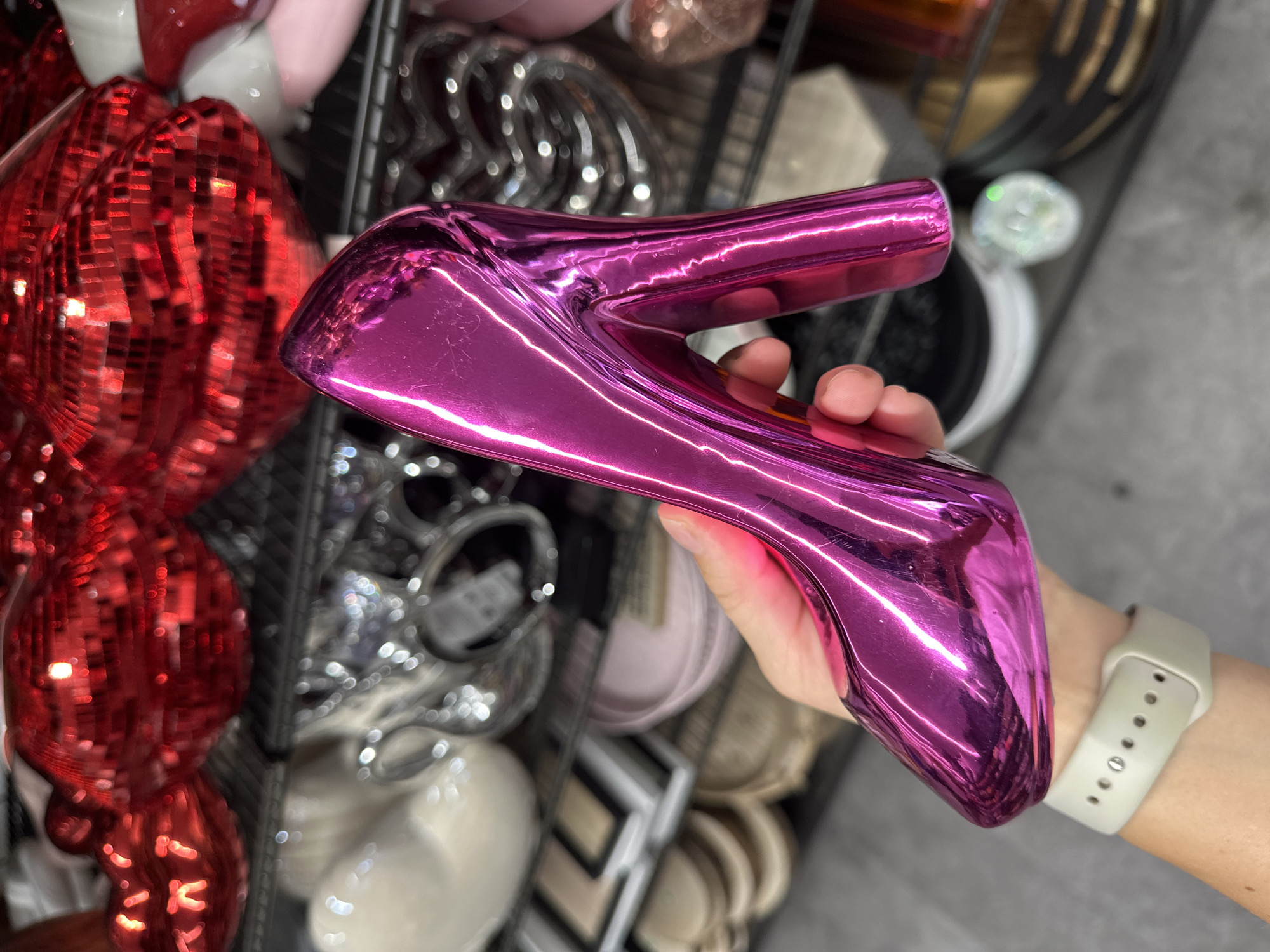 A close-up shot shows a person's hand holding a shiny, magenta-colored shoe figurine. The background is a display shelf filled with various decorative items, including red and silver objects. The hand holding the shoe has a light skin tone and a light-colored smart watch on the wrist. The setting appears to be a retail environment, possibly a home decor or gift shop, but without visual cues, definitively stating the location as Stade, Germany is not possible.