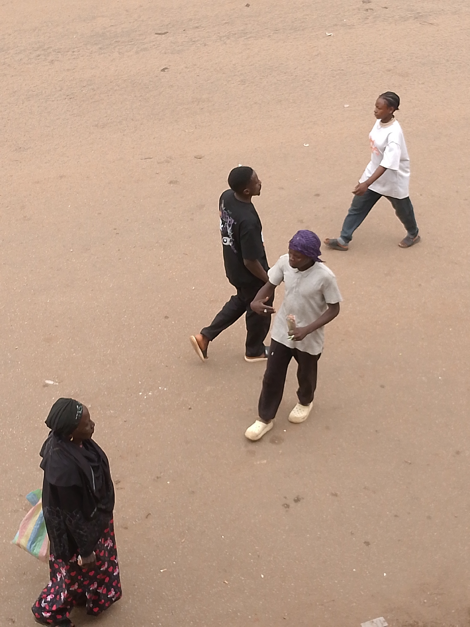 The image shows an elevated view of four people on a surface.
An adult female is standing on the left with a black head covering and black and patterned floral dress, carrying a striped tote bag.
An adult male is wearing a black t-shirt, black pants, and brown sandals, walking diagonally towards the upper right corner.
Another adult male is wearing a purple head wrap, grey t-shirt, black pants, and white Crocs, walking to the right while holding a clear bottle in his right hand.
A woman is wearing a white t-shirt, blue jeans, and brown shoes, walking to the upper right corner.
The surface is tan in color and has some variations in the shading.
The location is Bukuru, Nigeria.