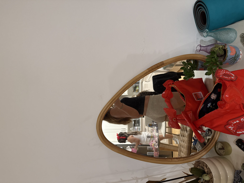 The image shows a teardrop-shaped mirror with a tan rattan frame. Reflected within the mirror are two women. The woman closest to the mirror is wearing a black top and light-colored shorts. In the background of the reflection is another woman. A glossy, red purse adorned with lip-shaped accents is positioned near the mirror's edge. Items like a yoga mat, small vases, and plants decorate the shelf above the mirror.