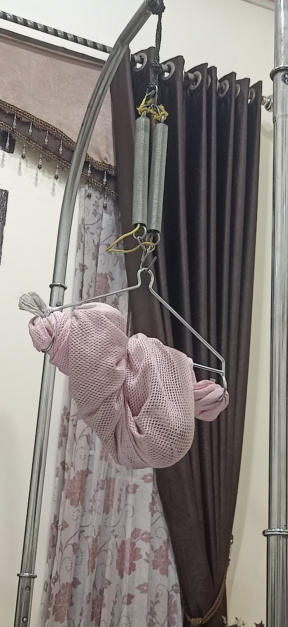 A pink mesh fabric bundle is suspended by a metal hanger from a set of springs attached to a curved metal stand. The stand is positioned in front of a window with two different curtains: a sheer floral-patterned curtain and a solid dark brown curtain. The scene appears to be indoors, likely in a residential setting in Lhokseumawe, Indonesia, given the context. The fabric bundle is bunched up and secured to the hanger by some white ties or cords.