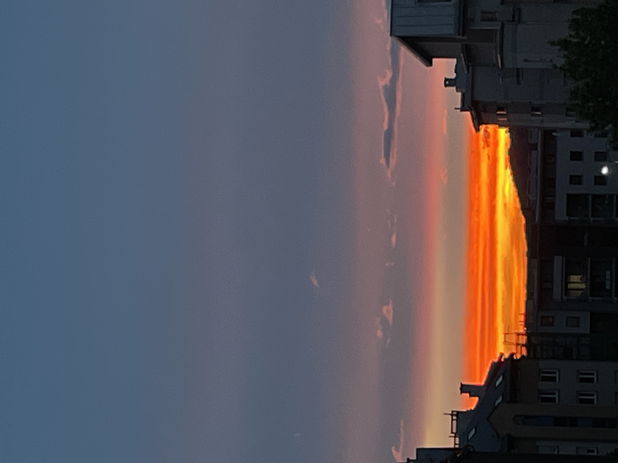 The image captures a vibrant sunset in an urban environment. The sky transitions from a dark blue on the left to a bright orange near the horizon. Buildings of varying heights are silhouetted against the sunset.
