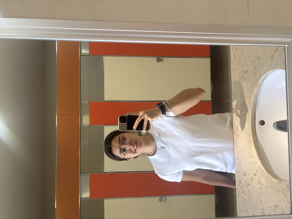 A mirror selfie of a young woman with dark hair and glasses wearing a white t-shirt. She is holding a black phone to take the picture. The reflection shows a public restroom with orange and beige dividers, a white sink, and a light-colored countertop. The mirror has a silver frame and reflects the brightly lit bathroom.