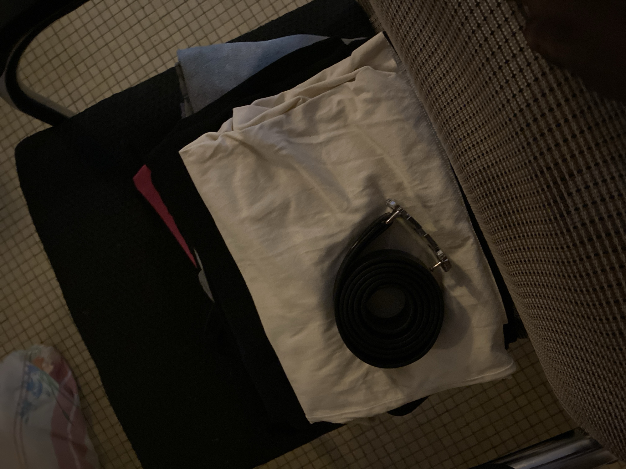 The image shows a black leather belt coiled on top of folded white fabric. The fabric is layered over other clothing items, including what appears to be black fabric and a pink garment. These items are arranged on a dark mat which is placed on a tiled floor. The lighting is dim, suggesting an indoor setting. The image is a close-up shot focused on the belt and fabric, with the background blurred. There is no indication of the city of Giginyu, Nigeria, in the image or its content.