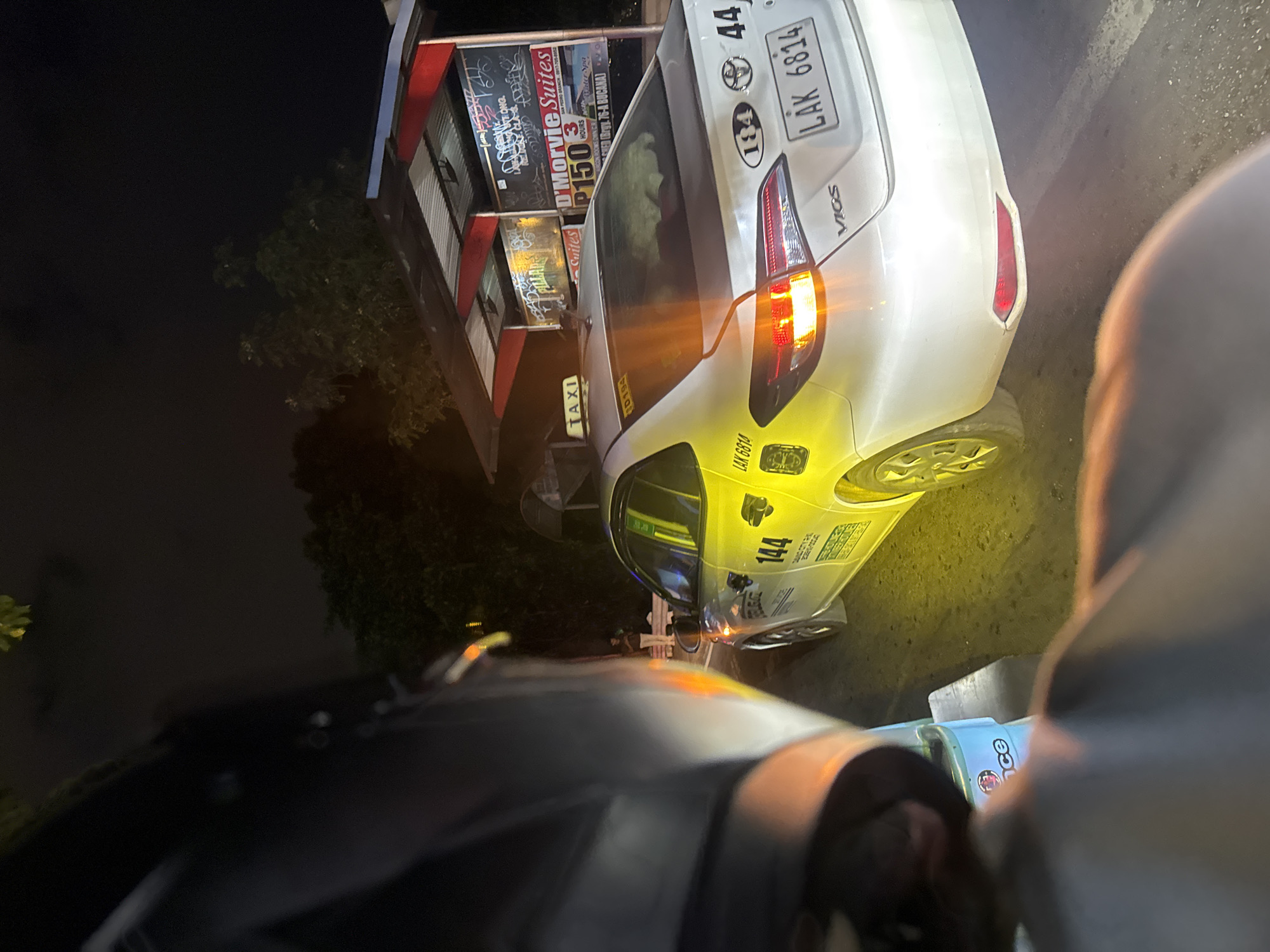 This image depicts a nighttime scene on a street in Davao City, Philippines, likely at a taxi stand. In the foreground, the left portion of a dark-colored motorcycle helmet is visible, suggesting the photo was taken by a motorcyclist. To the right, a white taxi is parked, its rear end angled towards the viewer. The taxi has yellow accents and markings, including the number "144" on its side and a "TAXI" sign on the roof. The license plate reads "LAK 6814". Behind the taxi, a small, covered shelter with red trim is visible, featuring advertisements for "D'Morvie Suites" and prices such as "P150 3 HOURS". The ground is asphalt, illuminated by the taxi's yellow headlights and taillights, creating a warm glow. A few trees are discernible in the background, silhouetted against the dark night sky. A person in light-colored clothing can be seen standing near the shelter, and another is visible in the distance. The overall atmosphere is that of a typical urban night scene, possibly at a pickup or drop-off point.