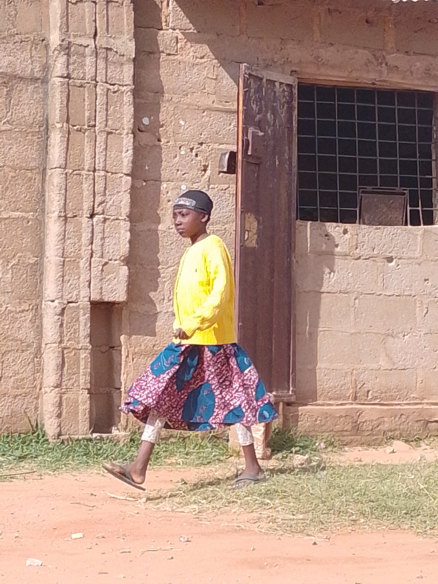 A person wearing a yellow long-sleeved shirt and a patterned skirt walks past a textured, light-colored brick building in Tumfure, Nigeria. The building has a dark metal door with peeling paint and a large window covered with a grid of dark bars. The ground is a mix of reddish-brown dirt and green grass. The person is wearing a dark headband and appears to be in motion, with their right foot off the ground, mid-stride. The lighting suggests it is daytime, with shadows cast on the building.