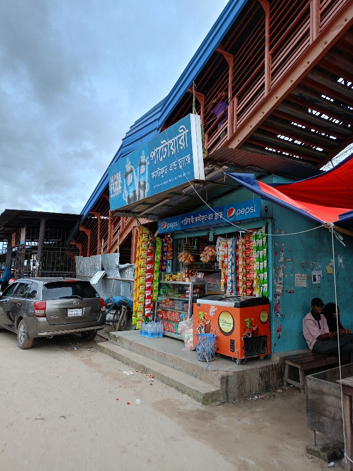 The image displays an outdoor street scene in Sujabad, Bangladesh, under an overcast sky. In the midground, a teal-colored shop is present, identified by a prominent blue and white sign above it that reads "পাটোয়ারী ফাস্টফুড এন্ড স্ন্যাক্স" (Patowari Fast Food & Snacks) in Bengali script, accompanied by Pepsi branding. A smaller Pepsi sign is also affixed to the shop's facade. The shop's open front reveals racks and display cases filled with packaged snacks and goods. An orange upright freezer with a circular yellow logo stands to the right of the shop entrance, alongside a blue plastic basket. Several large clear plastic water bottles are placed on the ground near the shop's threshold. A red and blue tarp awning extends from the shop's right side. To the left of the shop, a dark-colored SUV with a visible license plate reading "Dhaka Metro-G 34-2853" is parked on an unpaved dirt road. Two individuals are seated on a wooden bench to the far right of the shop; one male, wearing a light-colored shirt and dark cap, is partially visible. In the background, a large elevated structure with a blue corrugated metal roof and reddish-brown metal support beams is visible, appearing to be a multi-level pedestrian overpass or part of a station; one person in purple clothing is discernible on an upper level. The foreground consists of an unpaved dirt surface with some scattered debris.