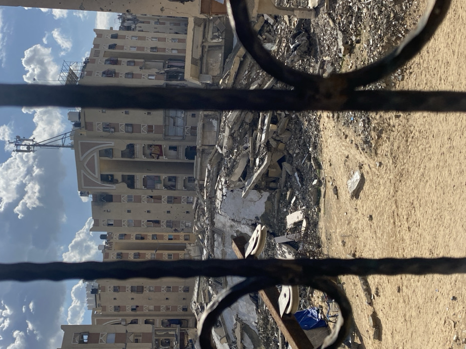 The image depicts a scene of extensive structural damage in Khan Yunis, Palestinian Territory, viewed through dark, ornate metal bars, possibly from a window or gate.
In the foreground, two dark, weathered vertical metal bars with some decorative curves are visible, partially obscuring the view. To the lower left, behind these bars, a blue tarp is present next to a rusty metal frame with two white pulleys. The immediate ground in front of the rubble consists of uneven sandy earth with scattered small debris.
The midground is dominated by large piles of fragmented concrete, twisted rebar, and shattered building materials, indicating the complete or partial collapse of structures. This debris field extends across the width of the image.
In the background, under a partly cloudy blue sky, stands a complex of multi-story, beige-colored residential buildings. These buildings exhibit widespread damage, including numerous projectile holes on their facades, shattered windows, and severely damaged or missing balconies. Some interiors are exposed through broken sections. The central building features prominent decorative arched elements on its upper floors and appears to be heavily scarred. A communication tower or antenna array is mounted on the roof of this central building, alongside multiple black water tanks. No people are visible in the scene. The overall environment suggests a conflict-affected urban area.