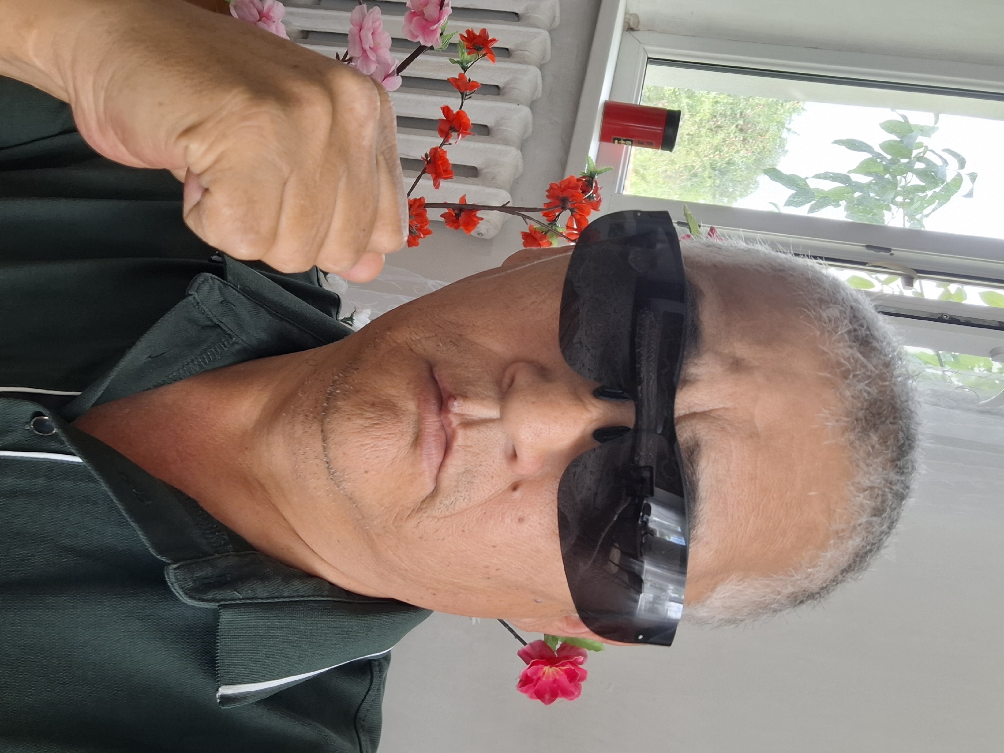 A close-up, waist-up, selfie-style photograph shows a middle-aged East Asian man with short, graying hair and a serious expression. He is wearing dark sunglasses that reflect the surrounding environment, a dark green collared shirt with white trim, and is holding his right fist up near his face. He has a small scar or mole above his upper lip. The man is positioned in front of a white radiator and a window. Behind the radiator are artificial branches with red and pink flowers. To the left of the man, on the windowsill, is a red cylindrical object. The background outside the window appears to be foliage and greenery. The overall impression is one of indoor setting, possibly a residential room, with decorative elements. The image does not provide specific details to confirm its location in Almaty, Kazakhstan.