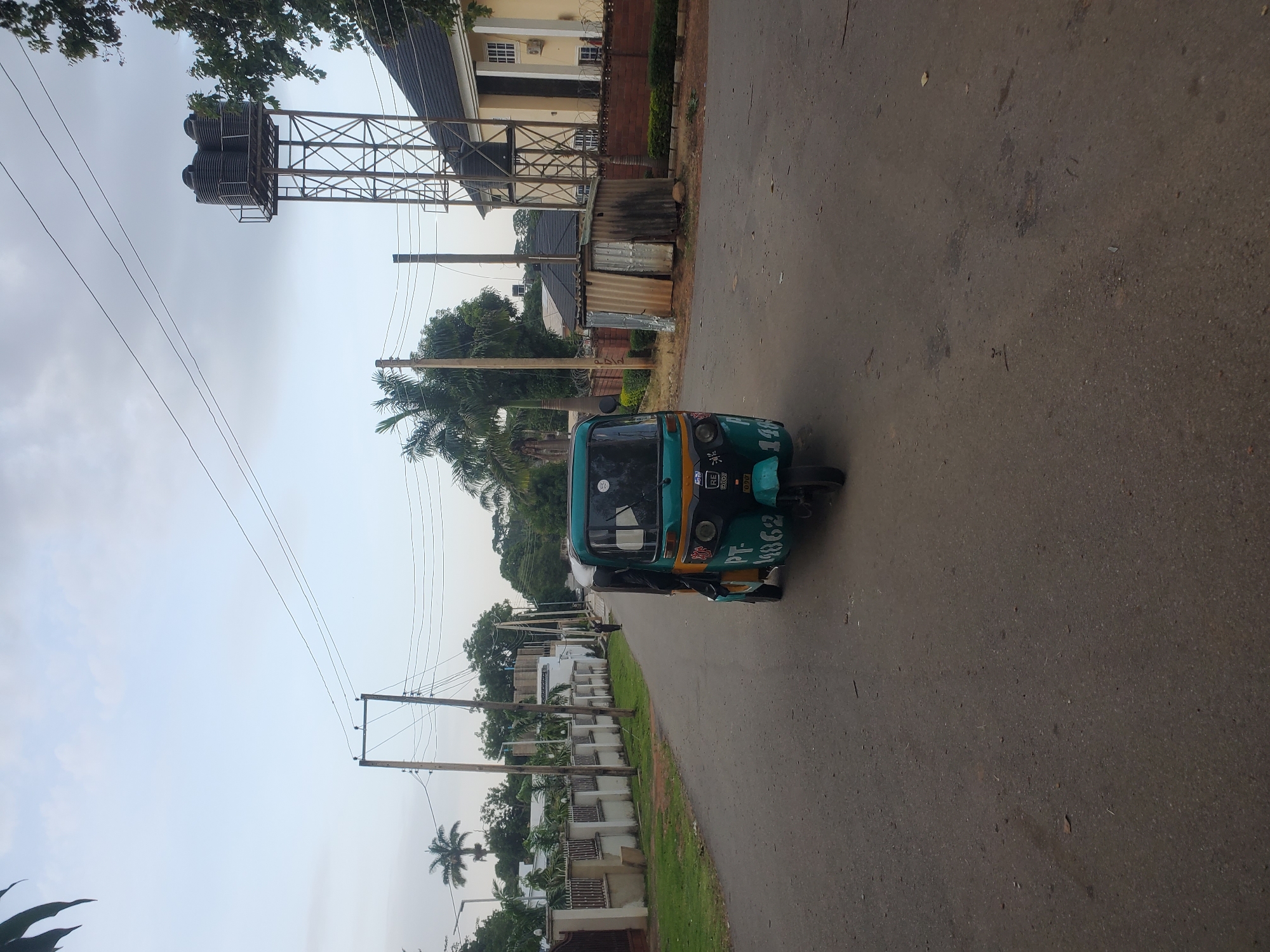 In Jos, Nigeria, an overcast sky hangs above a paved street. A three-wheeled auto-rickshaw, primarily green and yellow, occupies the center of the road, facing the viewer. Its license plate, partially obscured, reads "PT - 14862". A person can be seen partially visible inside the driver's compartment. The street is lined with a white fence with intermittent vertical posts and decorative railings, bordering a grassy area. Beyond the fence, palm trees and other foliage are visible, interspersed with modern houses, one prominently featuring a light yellow facade with white-trimmed windows and a dark roof. To the right, a tall metal water tower stands with multiple black water tanks atop it. A small, weathered corrugated metal structure sits near the base of the water tower. The ground is asphalt with scattered debris.