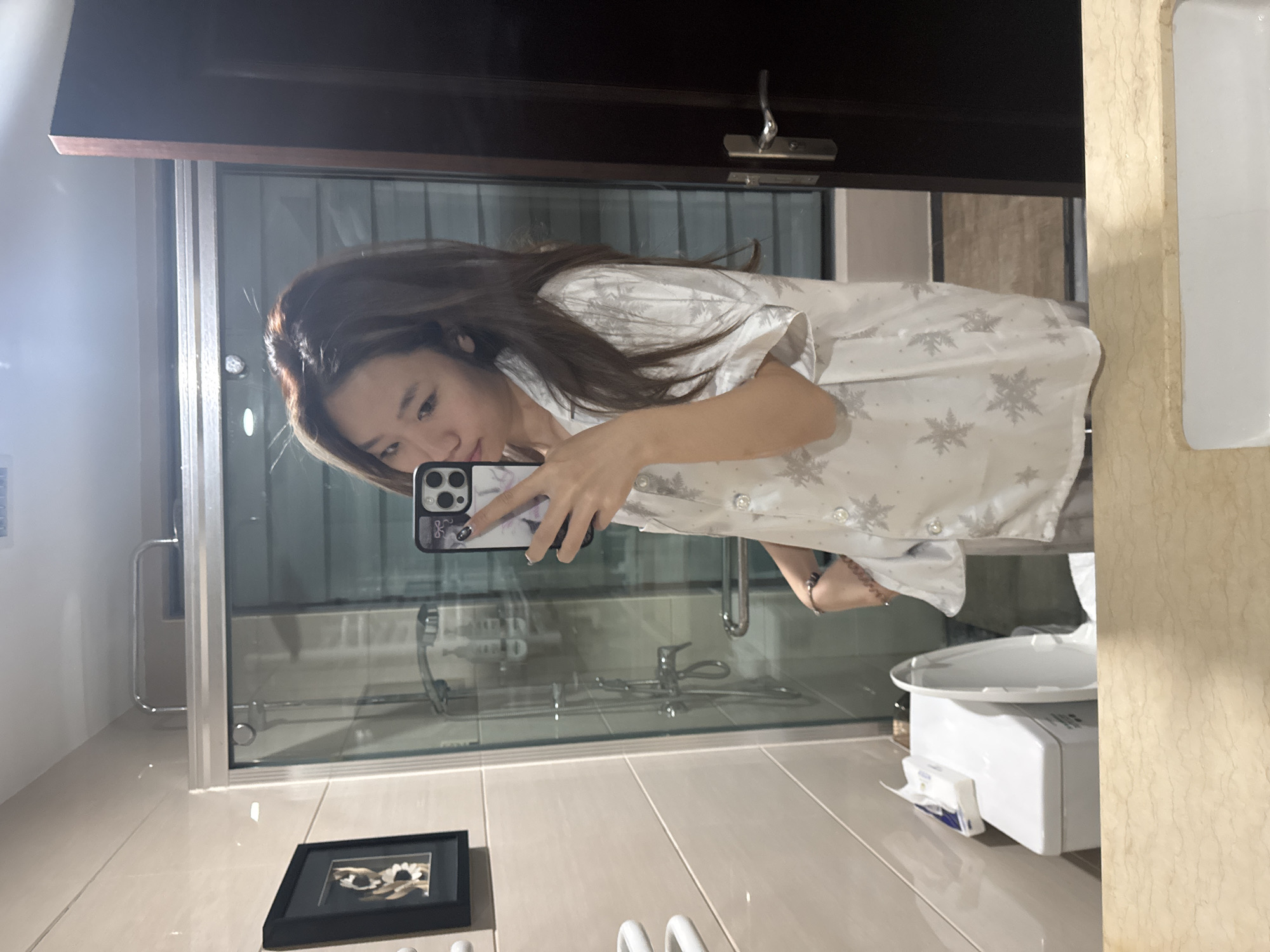 A young Asian woman is taking a selfie in a bathroom mirror. She has long brown hair and is wearing a white pajama top with a snowflake pattern. She is holding a smartphone with a white case in her left hand. The bathroom has a glass shower enclosure, a toilet with a white electronic bidet seat, and a framed picture on the floor. Based on the location metadata, the image was taken in Lanzhou, China.