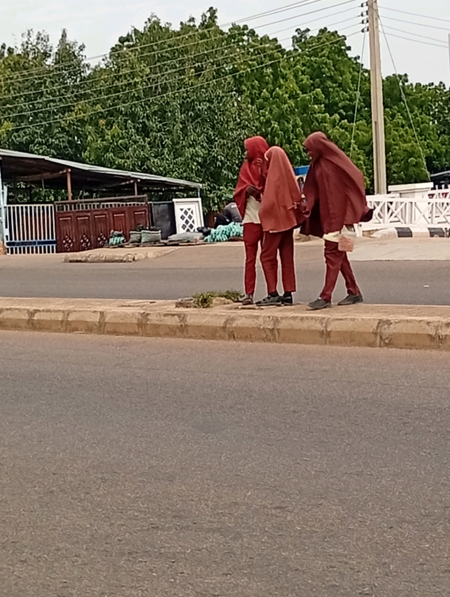 Three individuals wearing maroon hijabs and maroon attire are standing on a sidewalk next to a road. The individuals appear to be in a conversation or interaction. In the background, there are trees, a building with a covered roof, a fence, and a white decorative fence. Power lines are visible above the trees and a utility pole is present to the right of the frame. The ground in the foreground is a paved road. The scene appears to be in an urban or semi-urban environment in Tempure, Nigeria.
