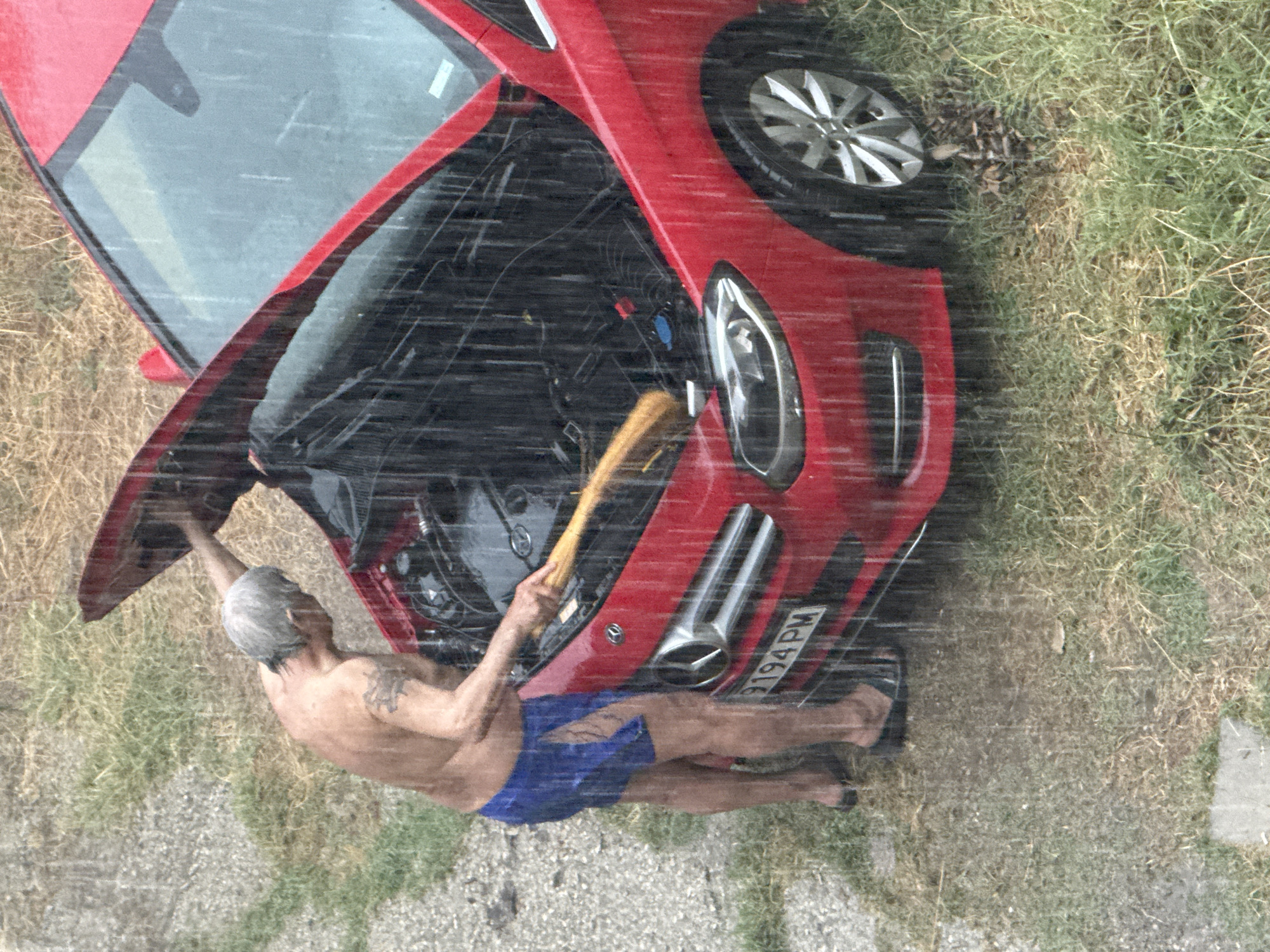 An shirtless man with gray hair, wearing blue shorts and dark flip-flops, is standing next to a red Mercedes-Benz car with its hood open. He is holding a yellow brush and appears to be cleaning or working on the car's engine. It is raining heavily, with visible raindrops impacting the scene. The car is parked on an uneven surface with a mix of gravel, grass, and some dirt. In the background, there is dry, yellowish grass. The license plate of the car reads "9194 PM". The scene appears to be outdoors, possibly in a residential or less maintained area, given the surrounding environment. This image is located in Burgas, Bulgaria.