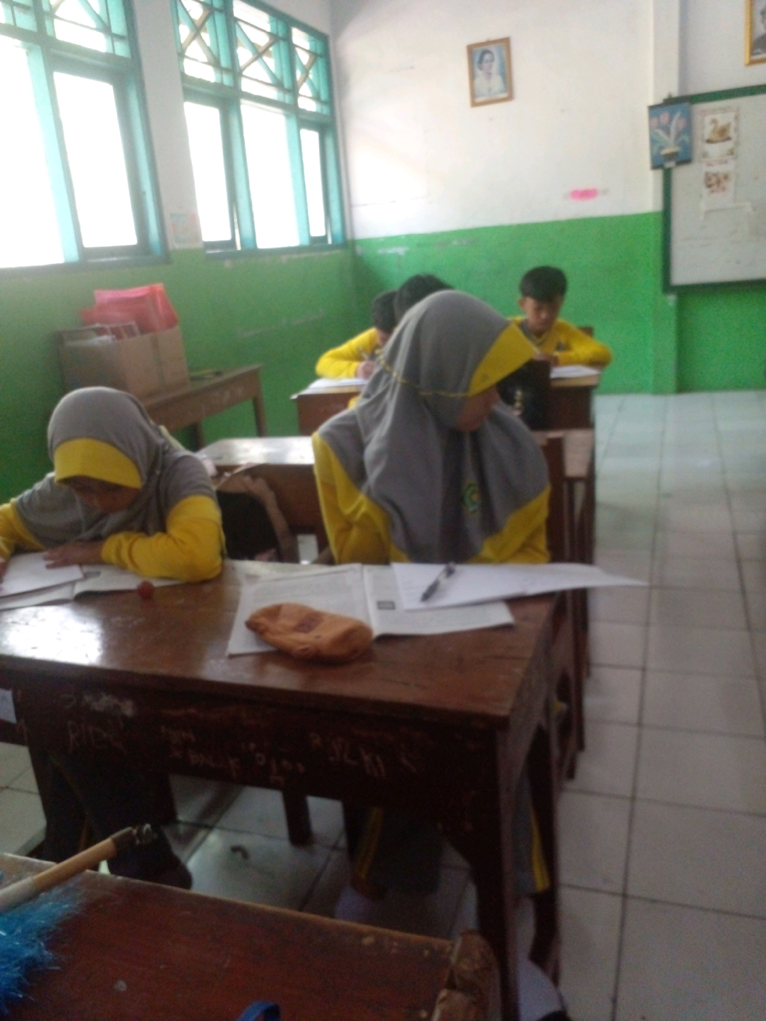 The image shows a classroom interior in Banyuputih, Indonesia. The scene depicts several students, identifiable by their yellow shirts and headscarves, seated at wooden desks and engaged in writing or reading. The desks are old and appear to have graffiti on them. In the foreground, a female student in a gray and yellow headscarf is focused on her work. The classroom has large windows with green frames that allow natural light to enter. The walls are painted green and white, and a framed picture of a woman hangs on the white wall. There is a whiteboard on the right side of the room with some writing and pictures on it. The floor is tiled. Other students and desks are visible in the background, suggesting a typical classroom setting.
