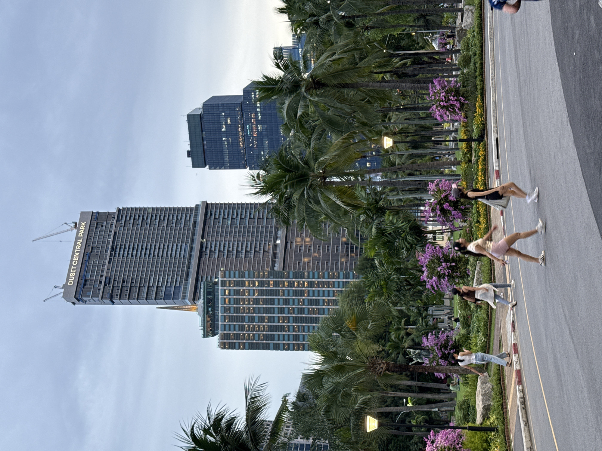 The image depicts a street scene in Bangkok, Thailand, featuring the "Dusit Central Park" high-rise building under construction. The street is lined with palm trees and vibrant purple and yellow flowering bushes. Four individuals are walking or running along the street, all oriented in the same direction, with two appearing to be engaged in physical activity. In the background, behind the palm trees, are additional high-rise buildings, illuminated with lights suggesting dusk or early evening. The sky is overcast.