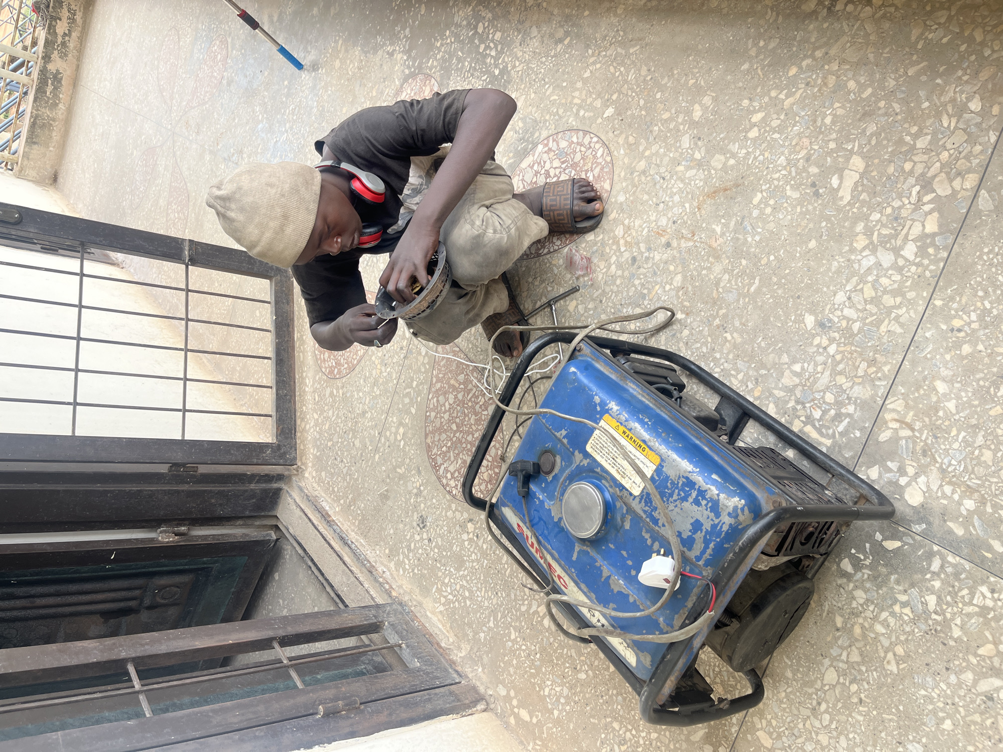 In the city of Gura Top, Nigeria, a young boy wearing a tan beanie and a black t-shirt is crouched on a terrazzo floor, working on a disassembled fan. He has red and white headphones around his neck. In front of him, to the left, is a blue generator with black metal framing. The background shows an open doorway with a metal gate and a patio area with metal railings. The floor has a distinctive speckled pattern.